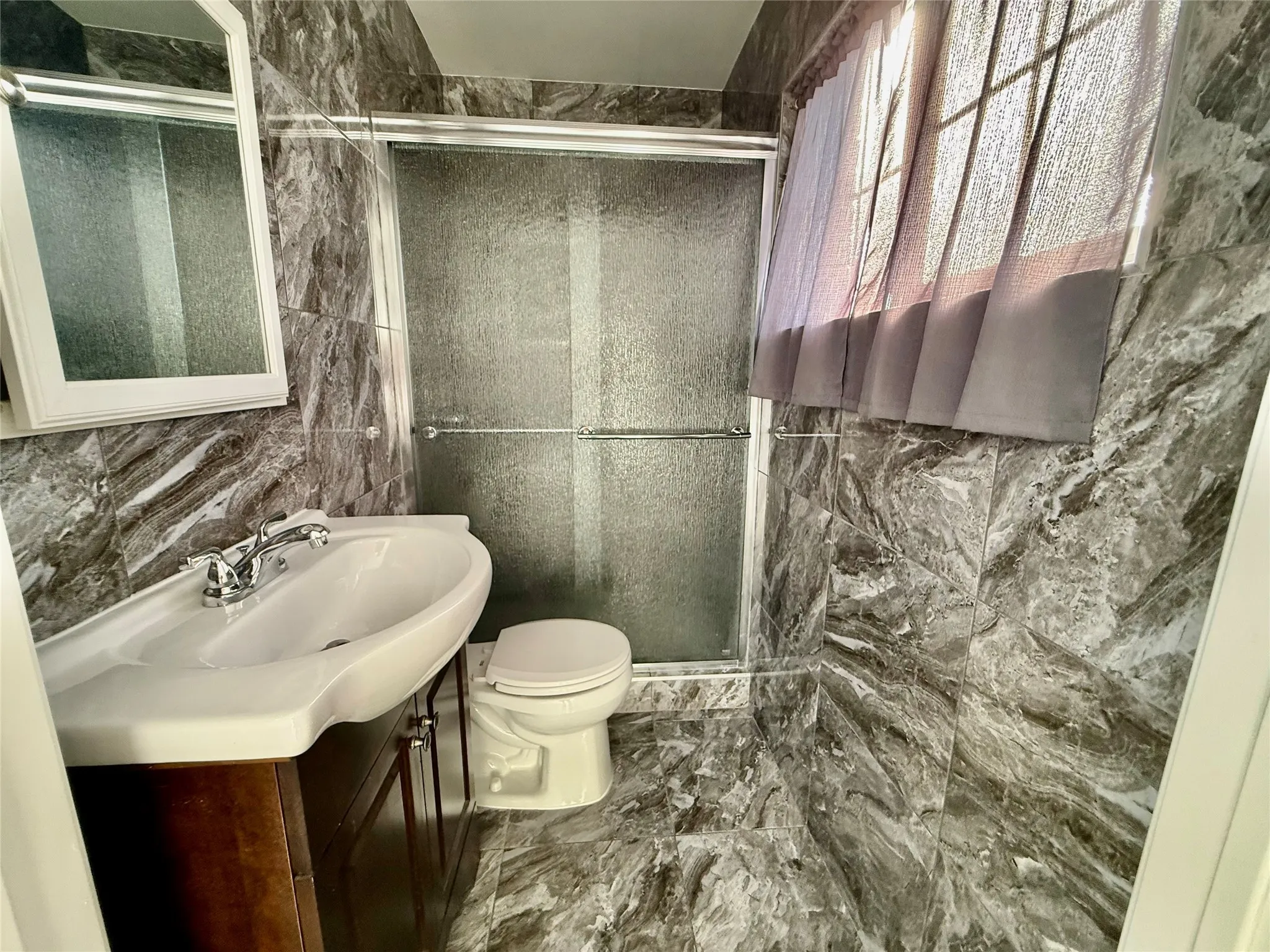 Tiled walls and flooring, wood-finish vanity with an integrated sink, polished chrome faucet, and a sliding glass shower enclosure