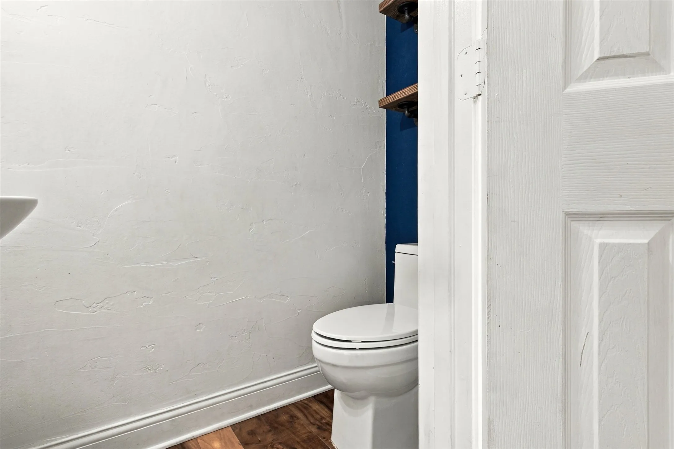 White pedestal sink, modern toilet, dark blue accent wall, wood-finish flooring, and white trim