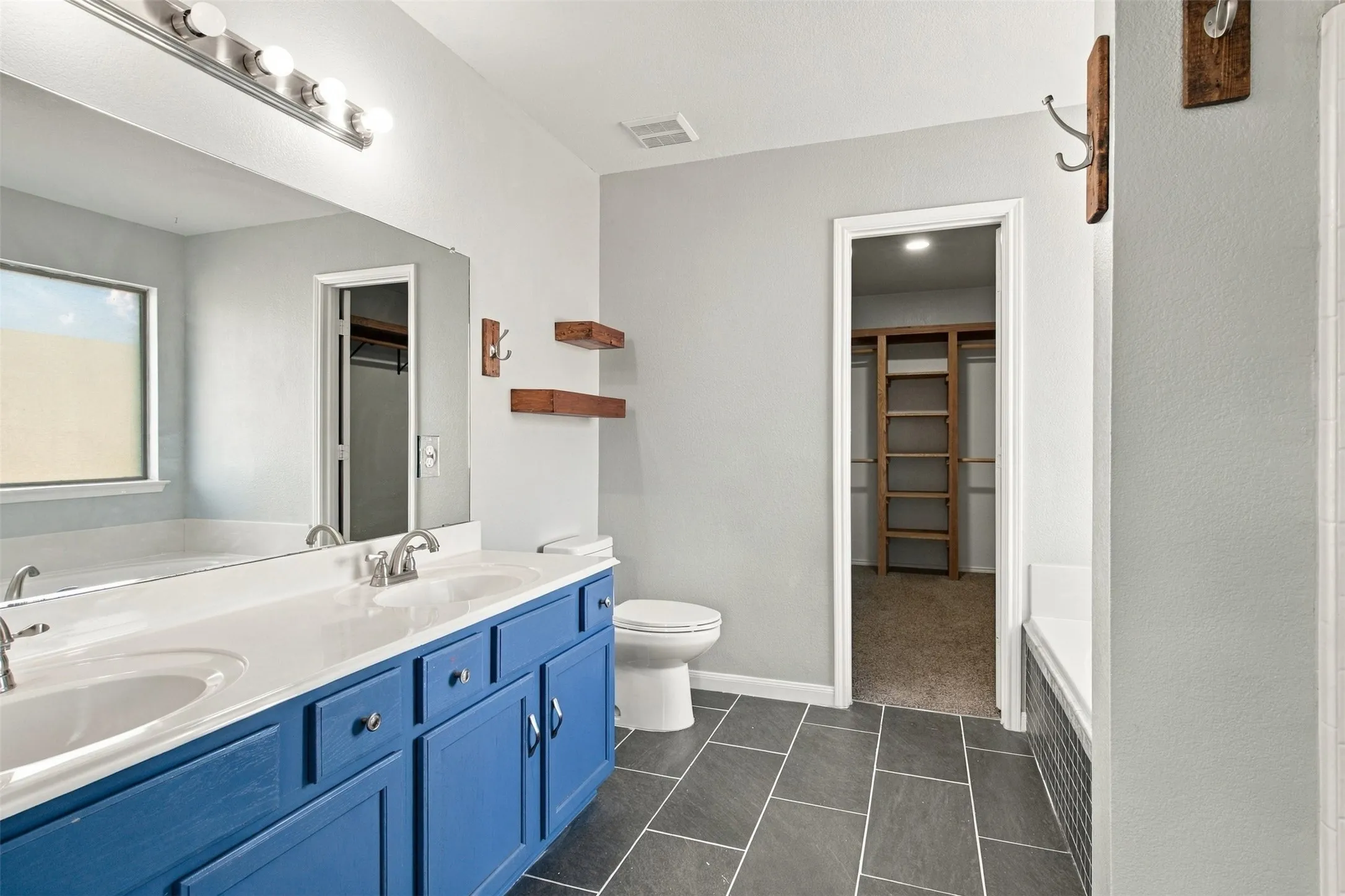 Dual sink vanity with blue cabinetry, full-width mirror, tile flooring, and built-in bathtub