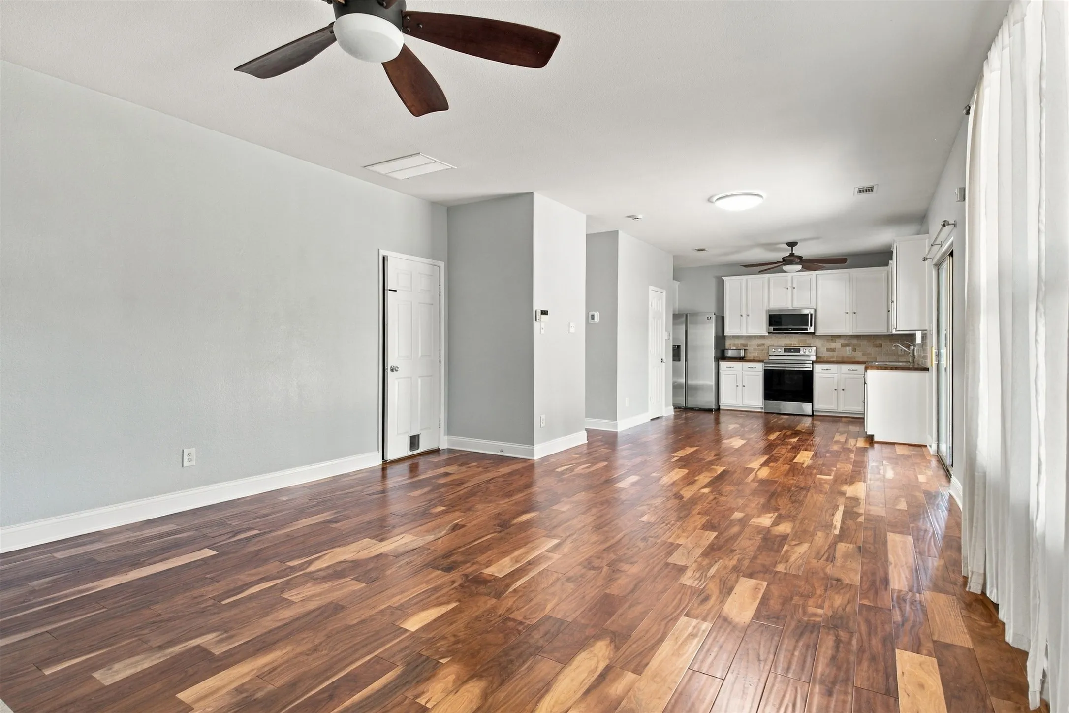 Open-concept living space with wood-finish flooring, ceiling fans, and a kitchen featuring white cabinetry, stainless steel appliances, and a tiled backsplash