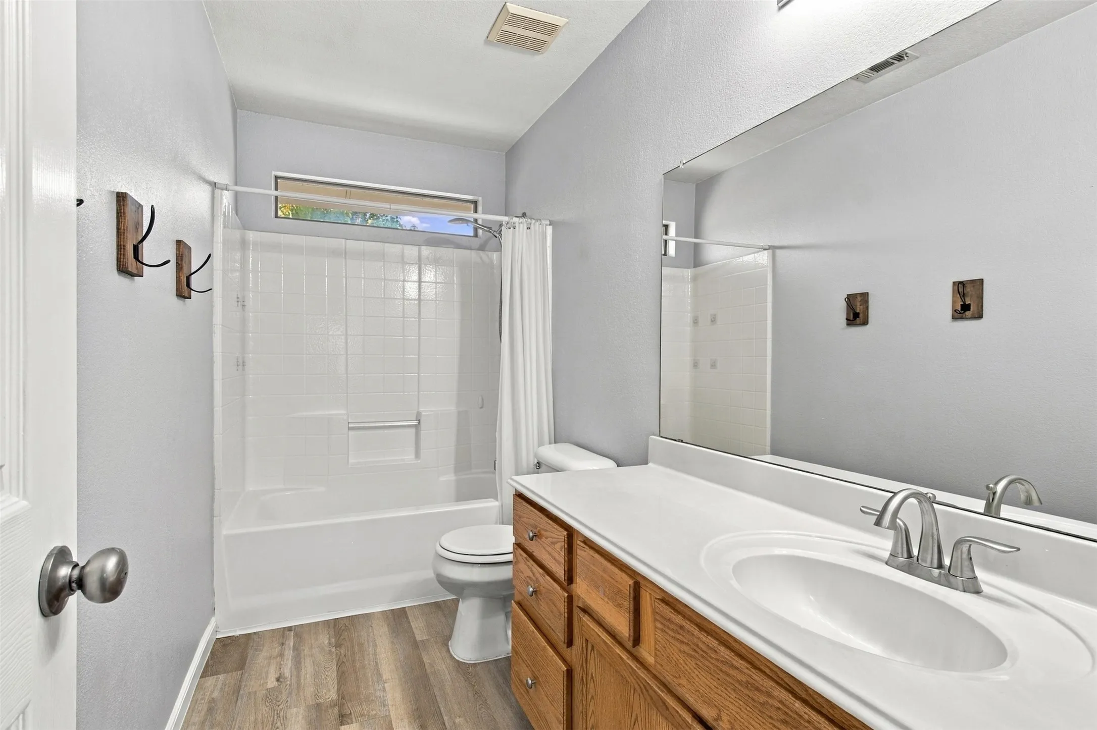 Bathroom featuring a combination shower and tub with white tile surround, wood-finish flooring, a long vanity with an integrated sink, a large mirror, and a horizontal window