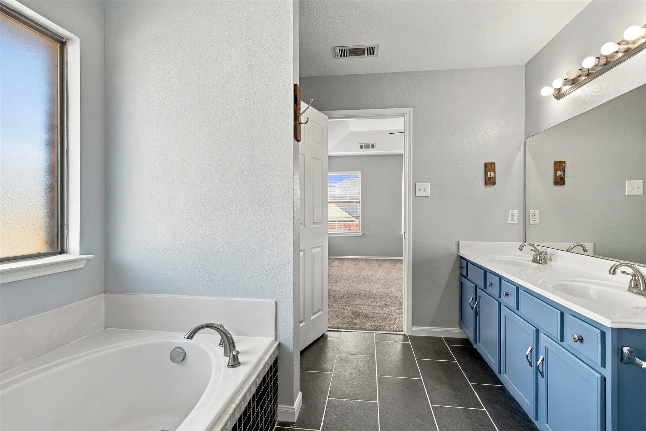 Double vanity with blue cabinetry, full-width mirror, tile flooring, and a built-in bathtub with tile surround