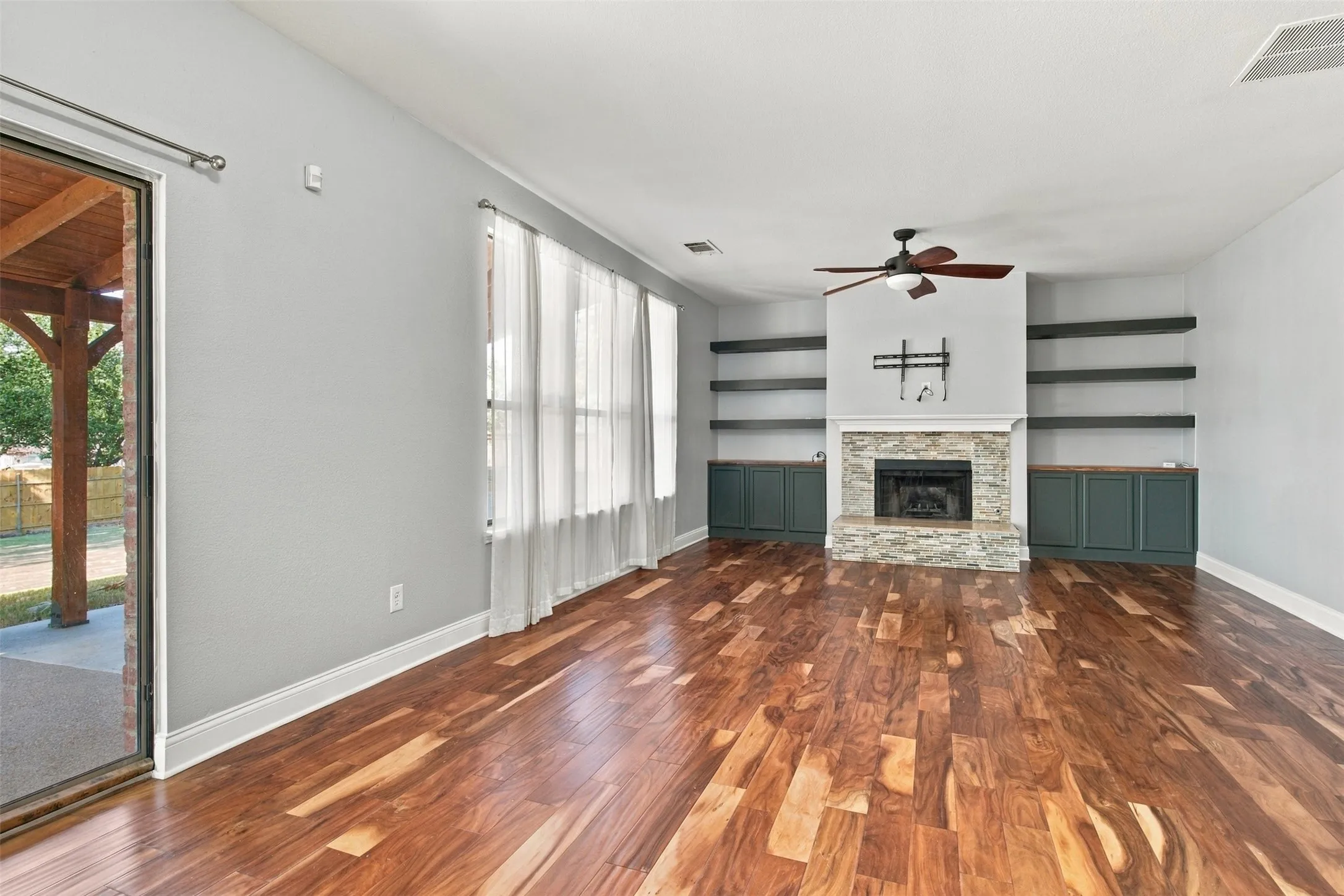 Wood-finish flooring, stone-clad fireplace with hearth, built-in shelving, integrated cabinetry, and ceiling fan