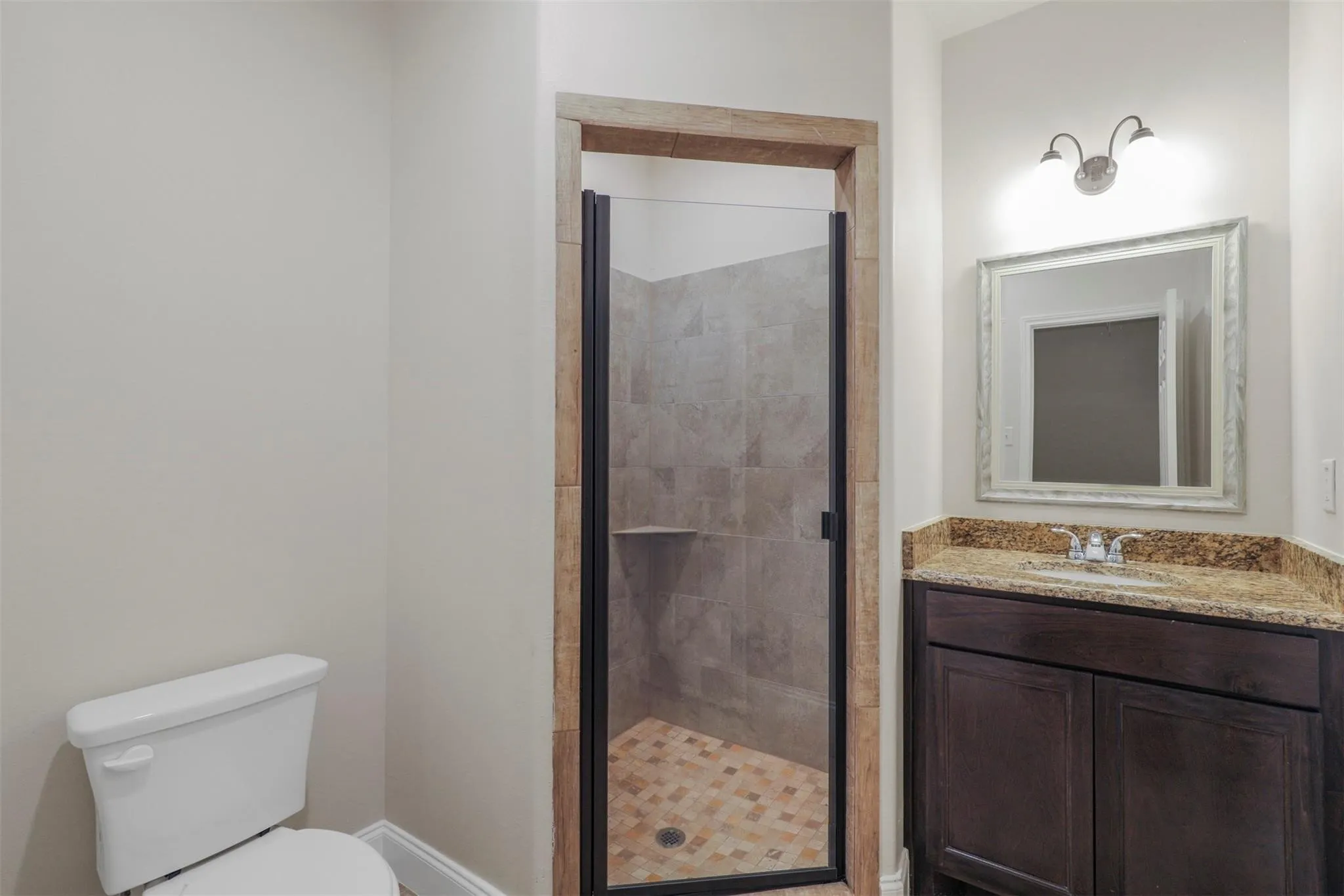 Shower with tiled surround and glass door, vanity with a stone countertop, built-in dark wood-finish cabinetry, and an ornate framed mirror