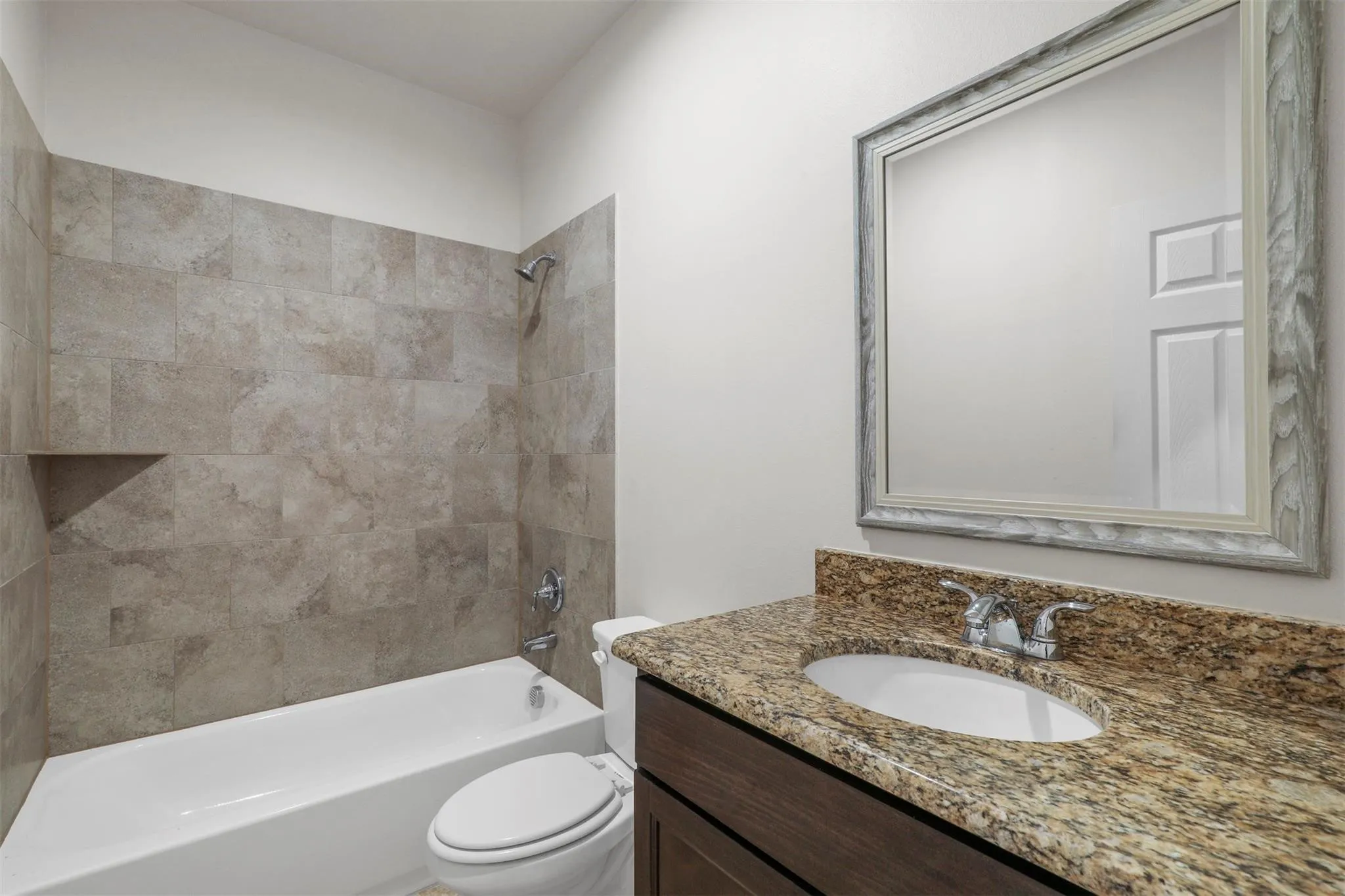 Bathroom featuring a tiled shower surround with a built-in niche, white bathtub, wood-finish vanity, stone countertop with an integrated sink, and a framed mirror