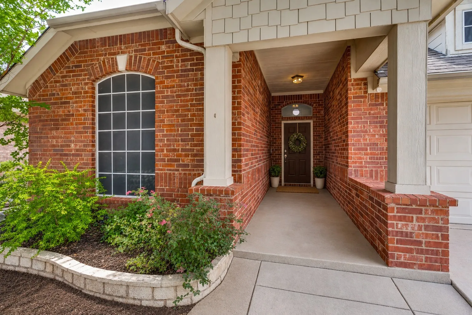Brick exterior with an arched window, covered entryway, concrete walkway, and a landscaped planting bed with stone retaining wall