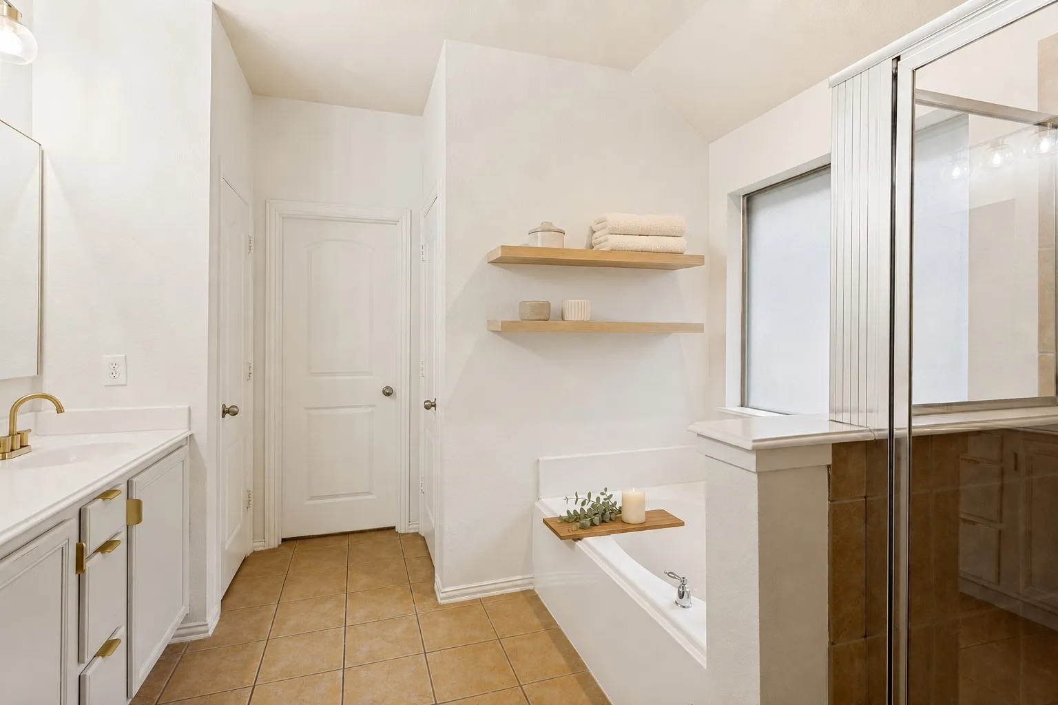 Bathroom featuring a soaking tub, glass-enclosed shower, dual vanity with brass-finish hardware, and floating shelves