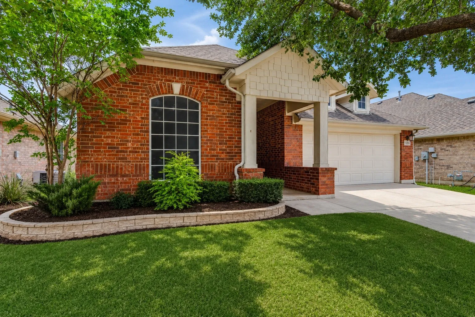 Brick exterior with an arched window, covered entryway, attached garage, and a tiered landscaping bed
