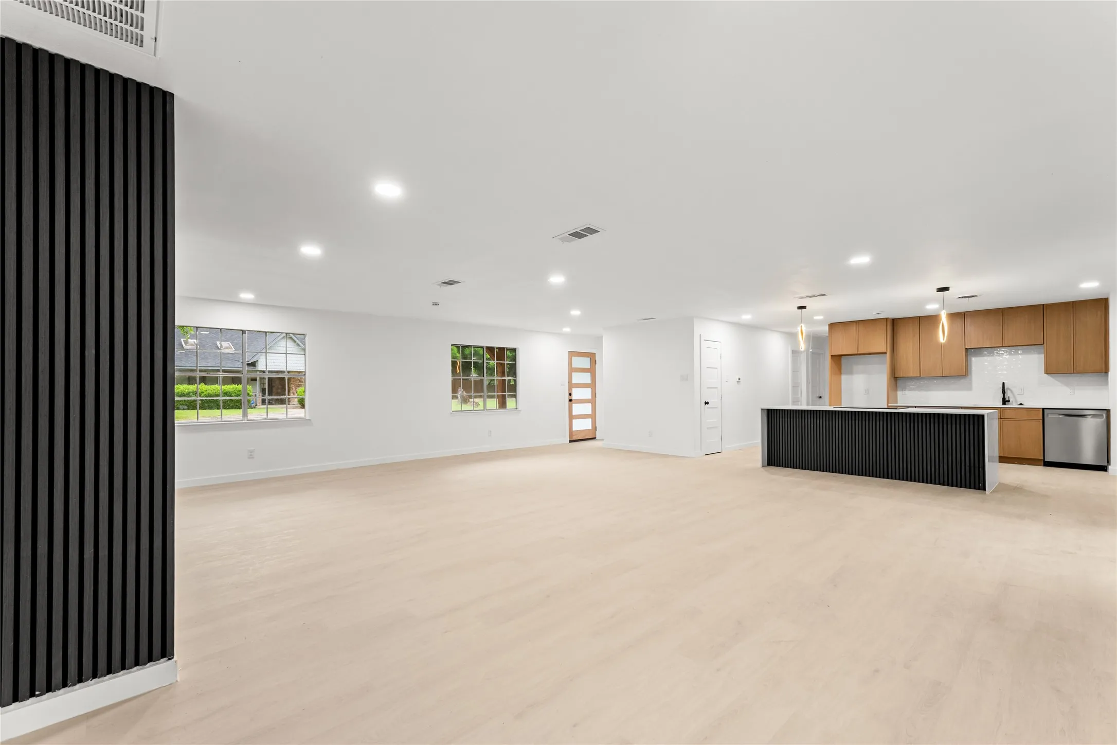 Open-concept living space with light wood-finish flooring, recessed lighting, and a modern kitchen featuring wood cabinetry, a central island with vertical paneling, and stainless steel appliances