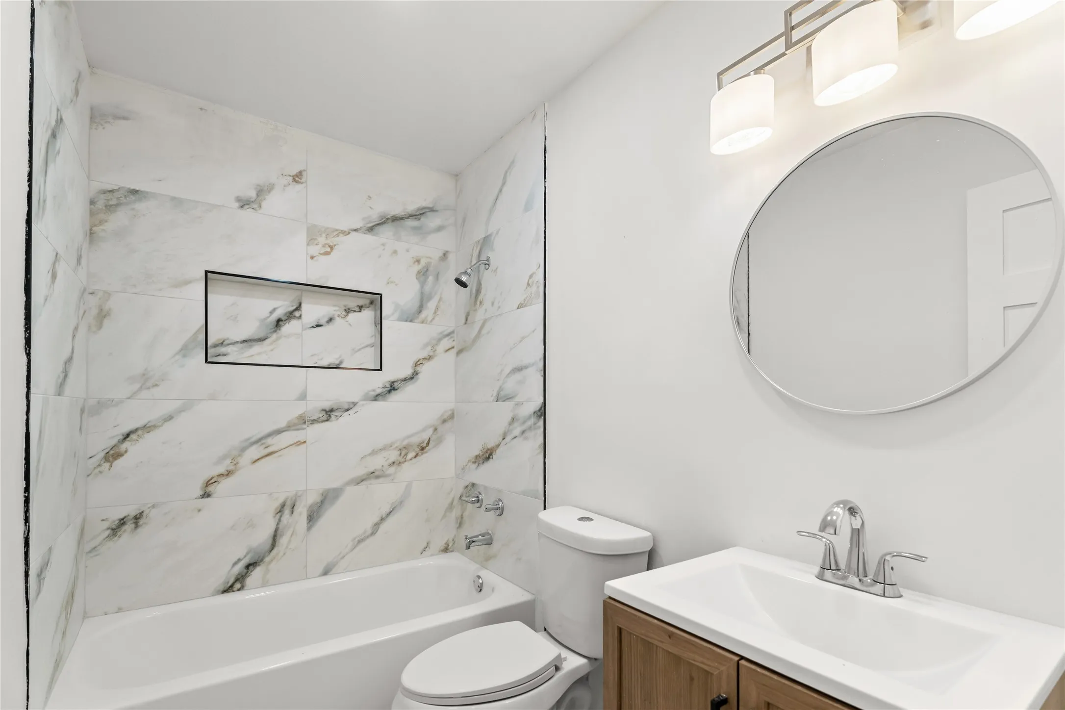 Bathtub with a tiled surround featuring an integrated shower niche, a wood-finish vanity with a white sink, and a circular wall-mounted mirror with an overhead light fixture