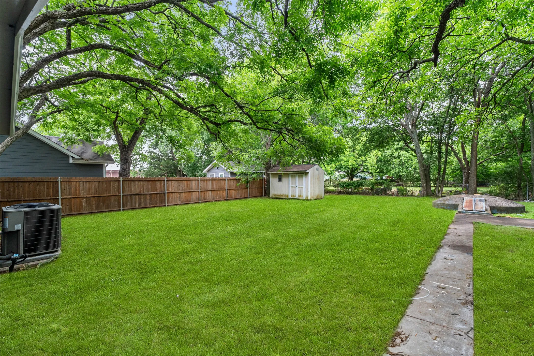 Expansive green lawn with mature trees, wood fence, and a storage shed