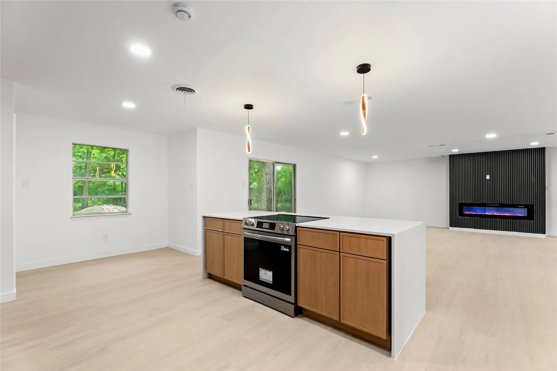 Open-concept living space with light wood-finish flooring, a kitchen island featuring wood cabinetry and a cooktop, recessed lighting, and a linear electric fireplace set into a textured accent wall
