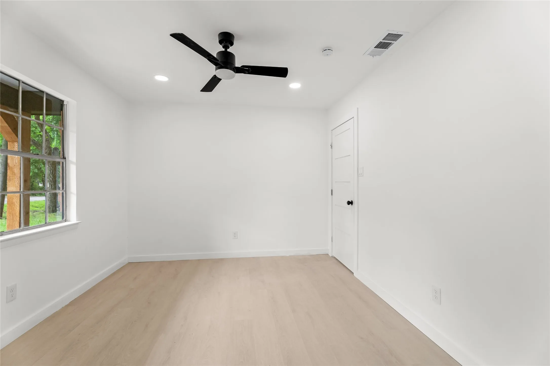 Light wood-finish flooring, ceiling fan with integrated lighting, recessed lighting, white panel door, and a large window