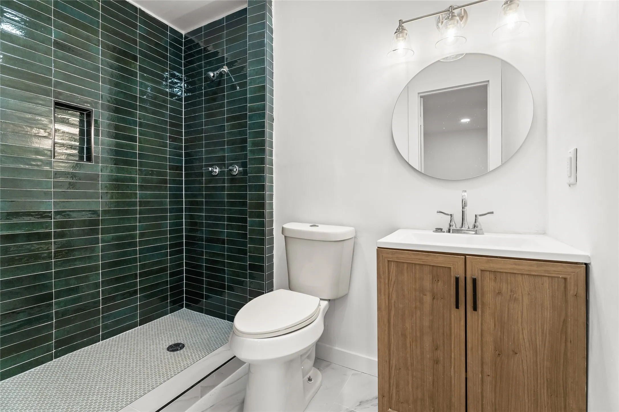 Walk-in shower with dark green subway tile, integrated shower niche, white hexagonal mosaic shower pan, light wood-finish vanity with a white countertop, and a circular mirror