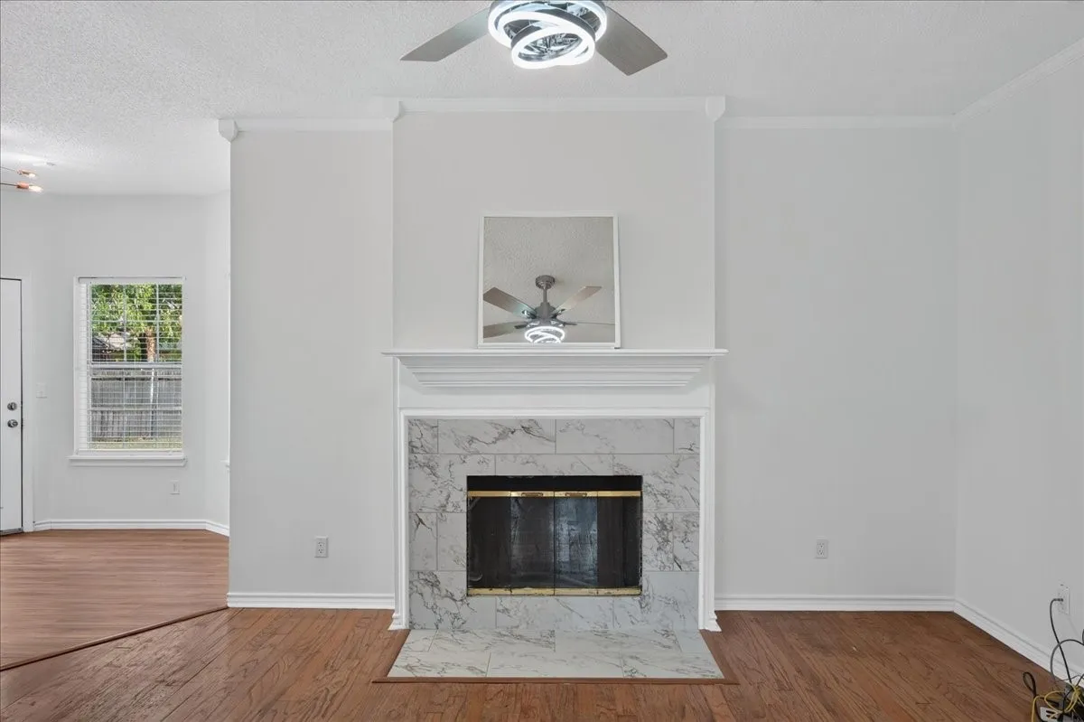Wood-finish flooring, white fireplace mantel, fireplace with stone surround, ceiling fan with integrated lighting, crown molding