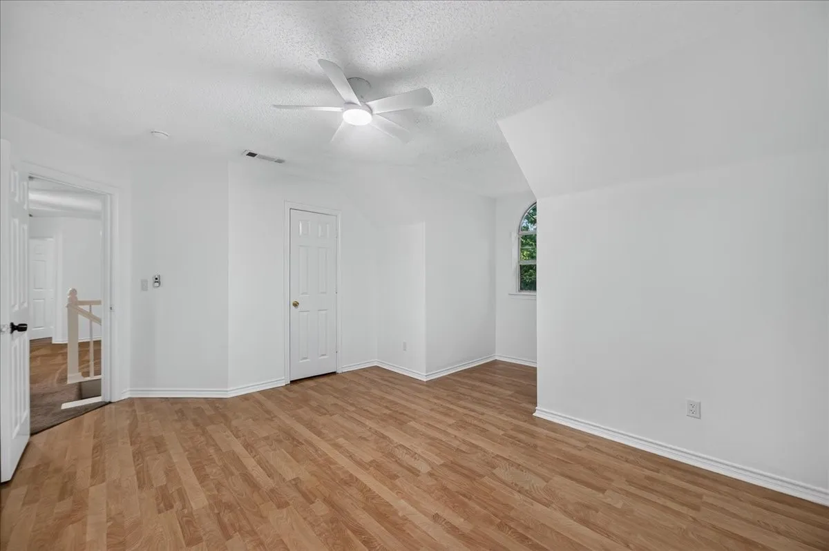 Wood-finish flooring, white walls, angled ceiling, built-in ceiling fan with light, and an arched window