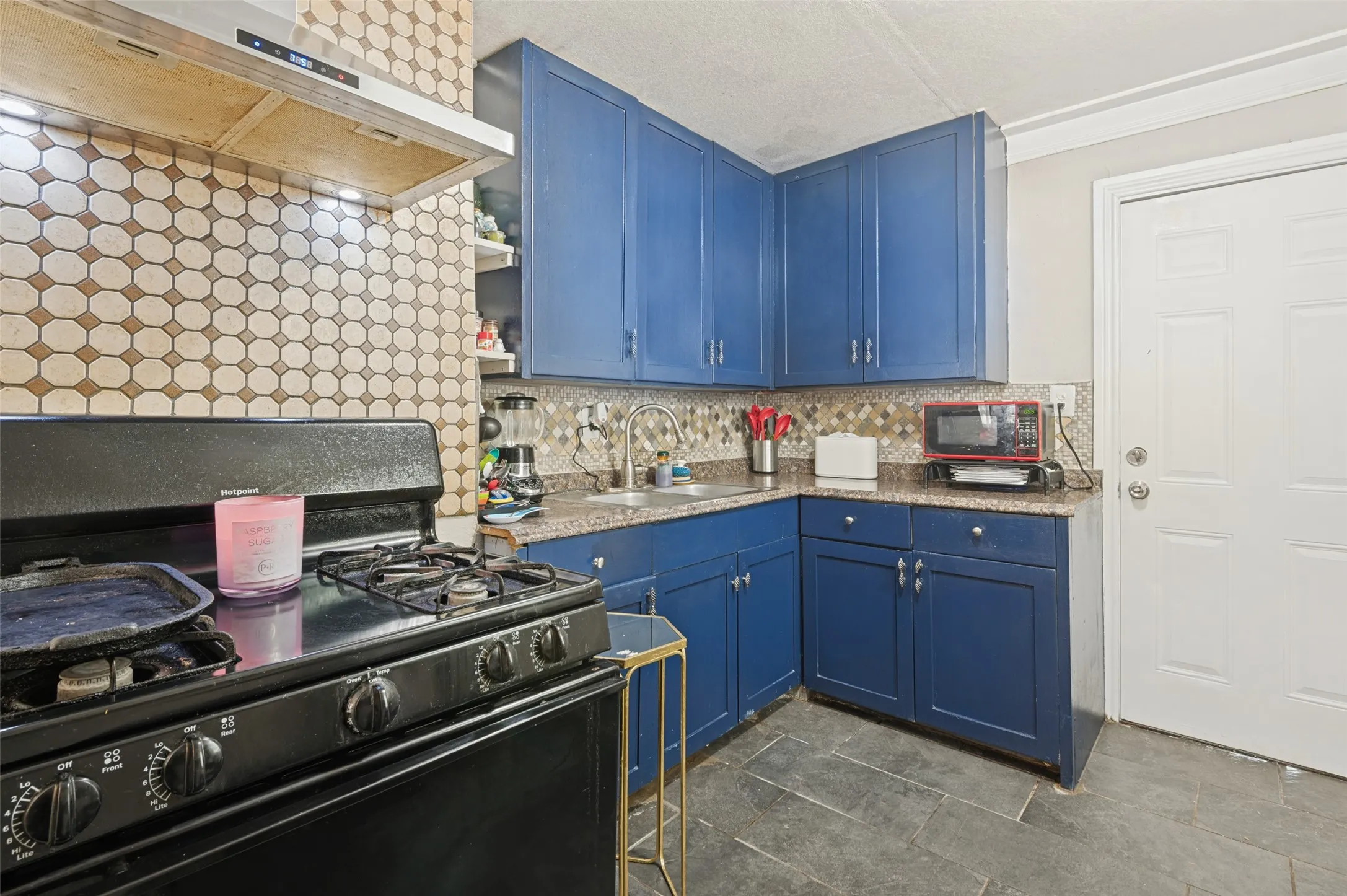 Blue cabinetry, patterned tile backsplash, dark stone-finish countertops, stainless steel range hood, and dark tile flooring