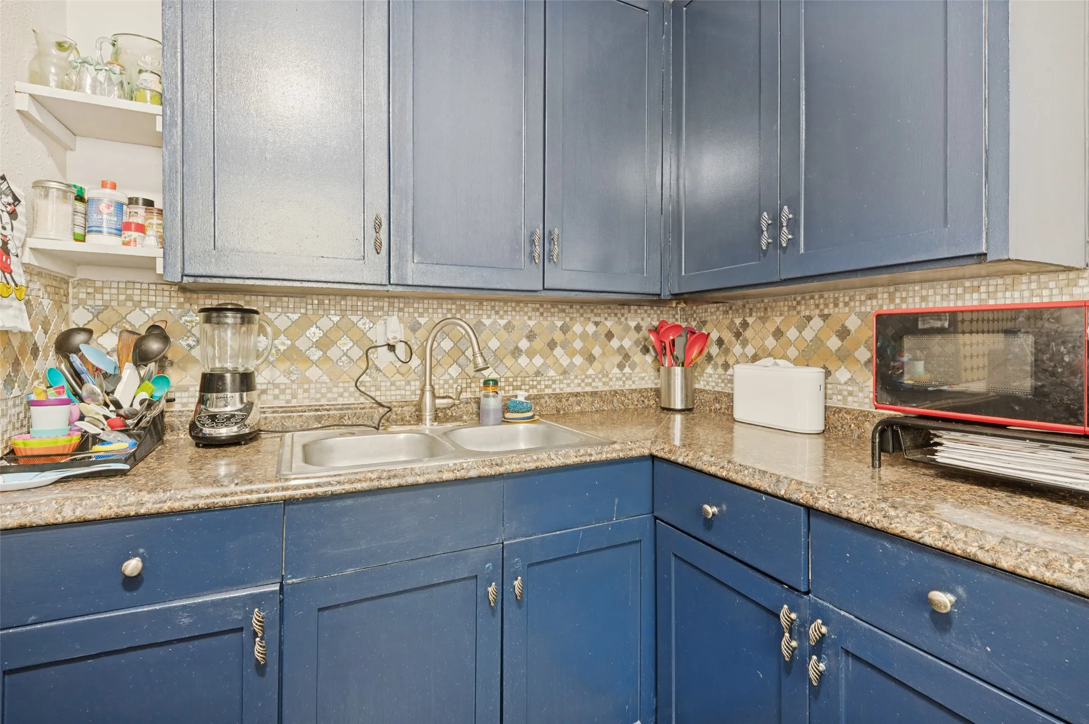 Blue cabinetry with varied hardware, dual basin stainless steel sink, mosaic tile backsplash, and speckled countertops