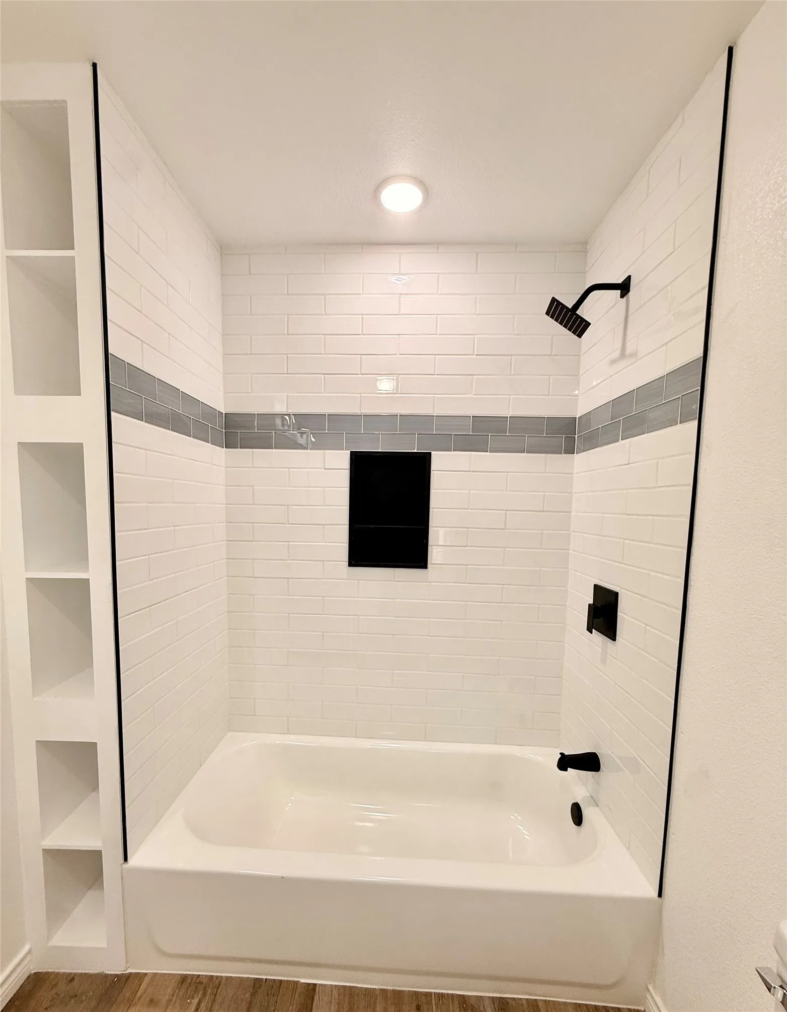 Bathtub with white subway tile surround, matte black fixtures, built-in shelving, and a recessed shower niche
