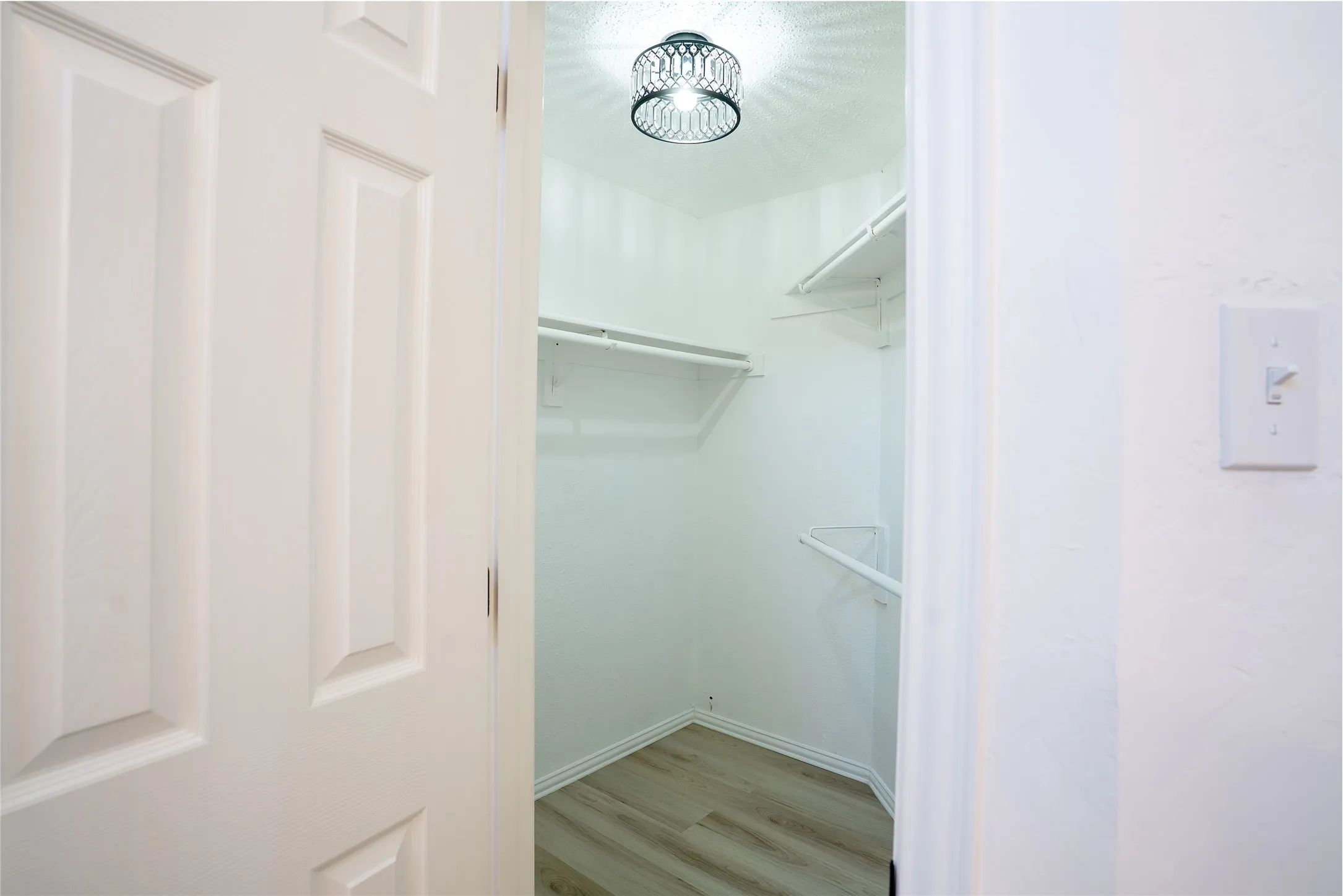 White paneled door opening to a closet with wood-finish flooring, built-in shelving, and a decorative ceiling light fixture