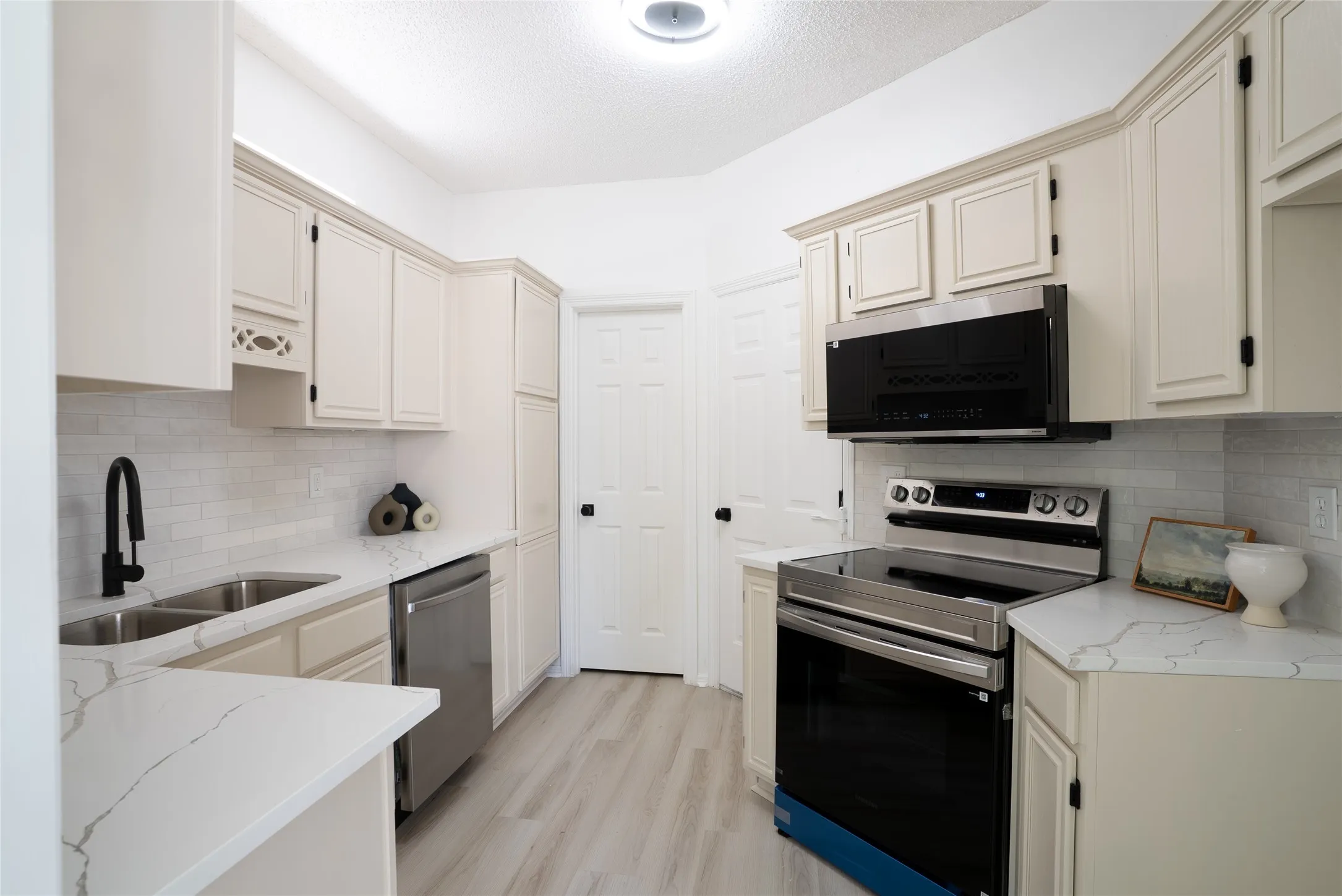 Kitchen featuring light-toned cabinetry, light-toned countertops, a stainless steel double sink with a black gooseneck faucet, a stainless steel dishwasher, and a stainless steel range with an overhead microwave