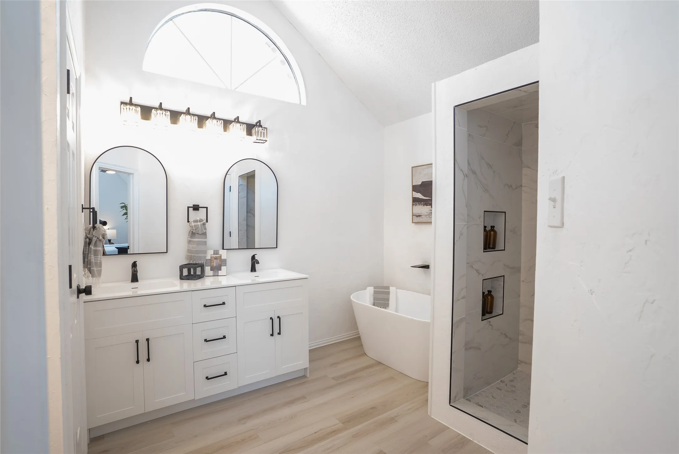 Arched window with gridded design, dual vanity with white cabinetry and black hardware, freestanding bathtub, and a glass-enclosed shower with tiled walls and recessed shelving