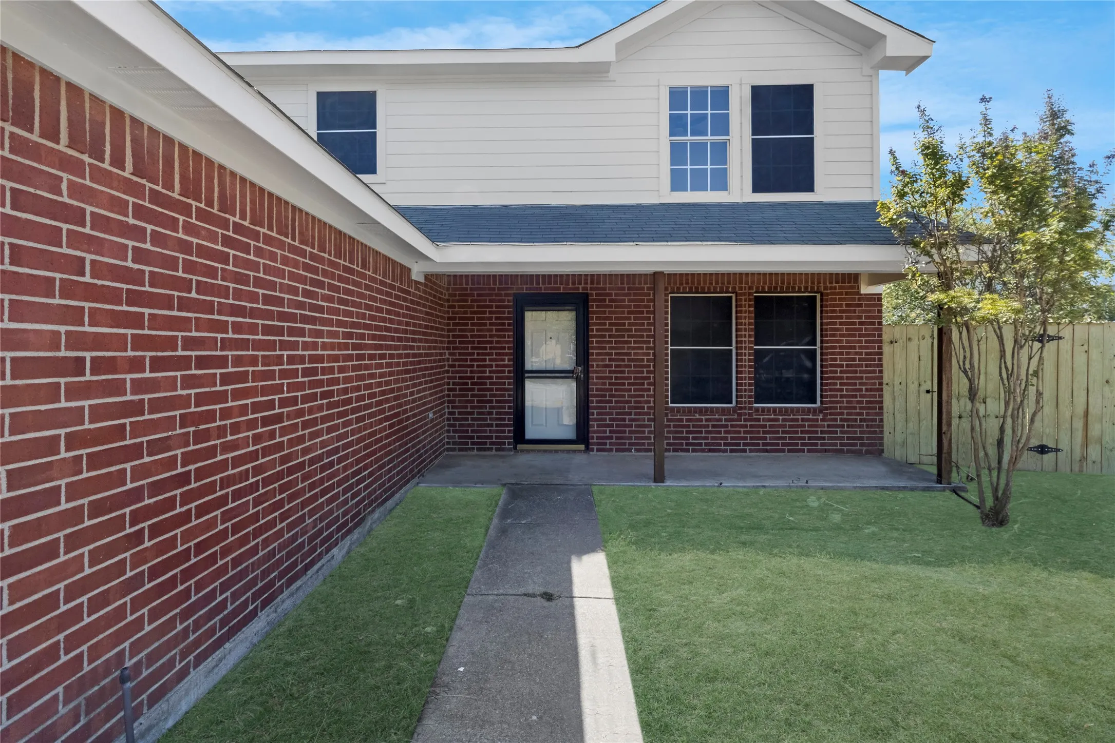 Brick and siding exterior with a covered porch, concrete walkway, and a wooden privacy fence