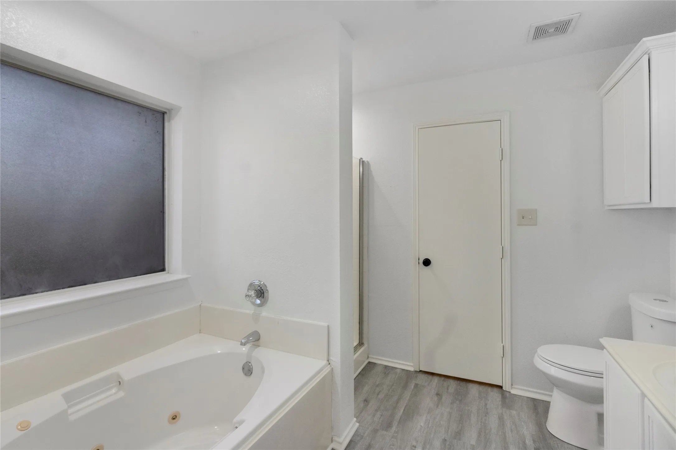 White jetted tub with chrome fixtures, frosted window, and wood-finish flooring