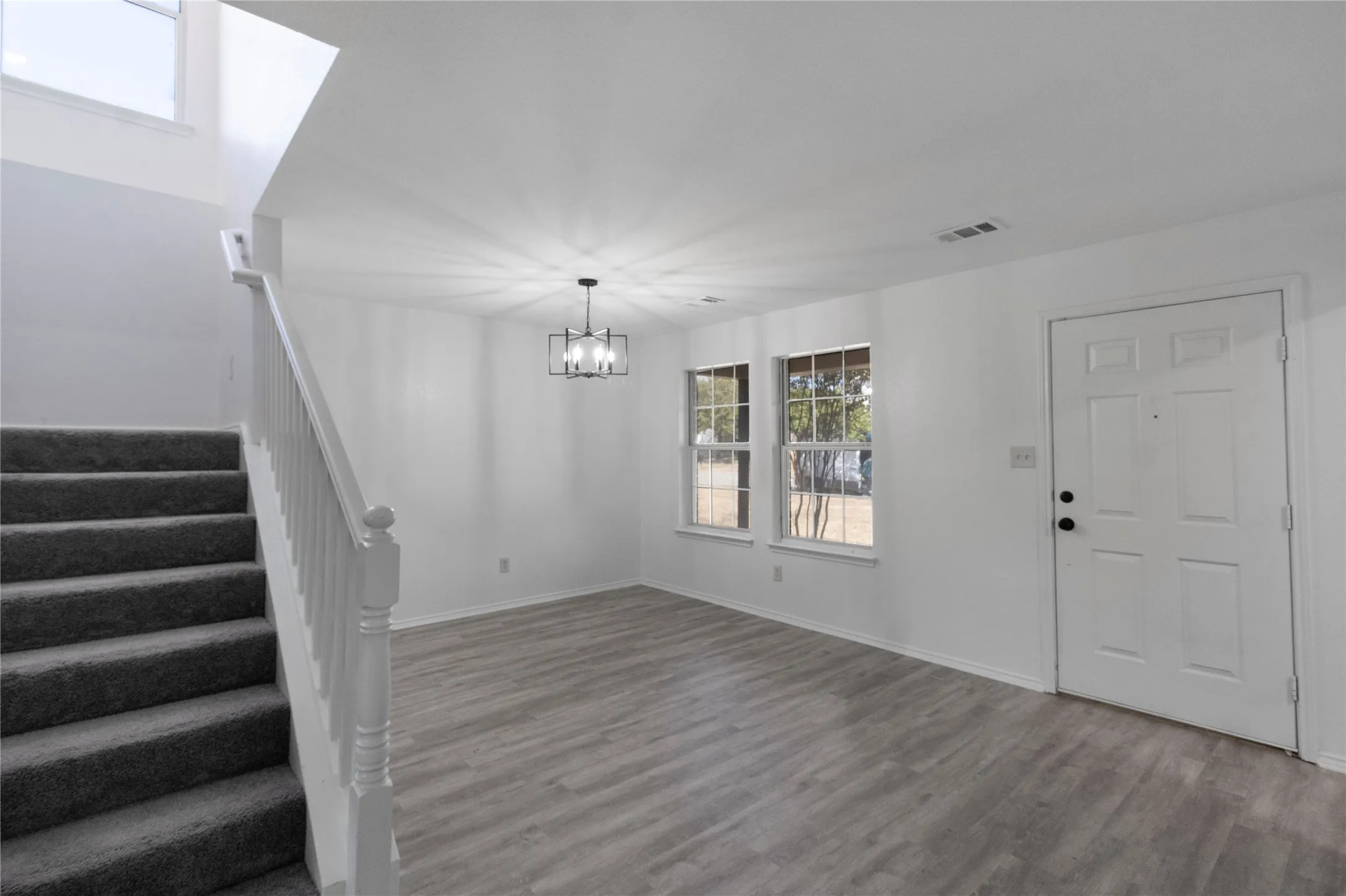 Wood-finish flooring, staircase with carpeted treads and white handrail, two windows, and a modern chandelier