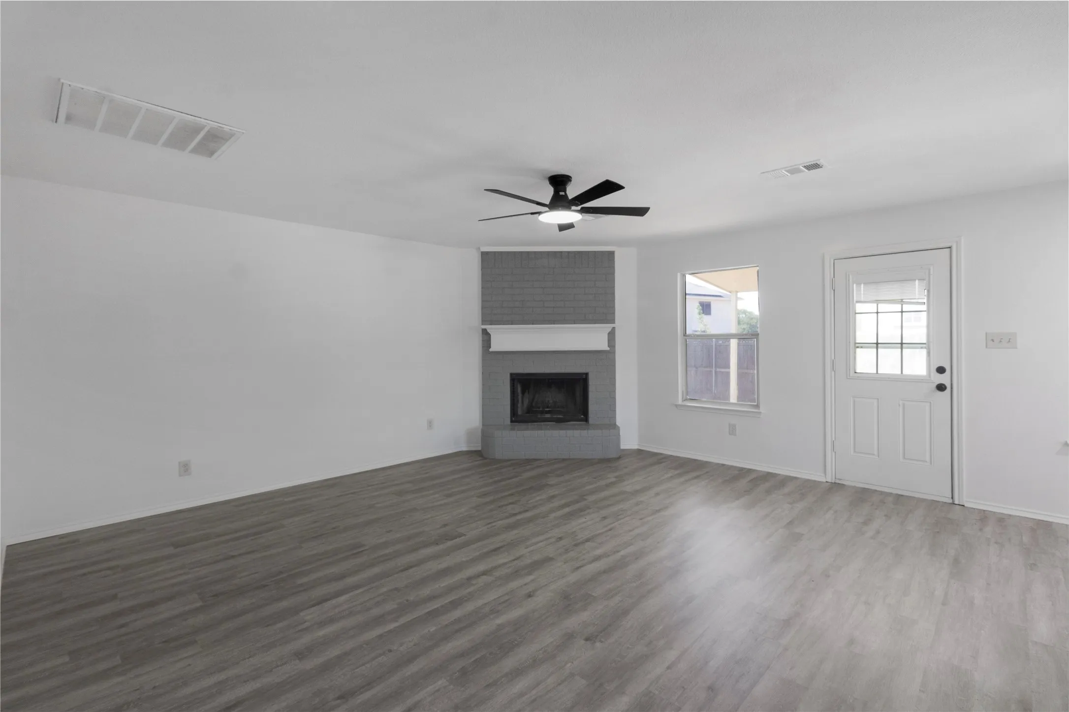 Wood-finish flooring, brick-faced fireplace with mantle, ceiling fan with integrated lighting, rear exterior door with glass inserts, and a window providing natural illumination