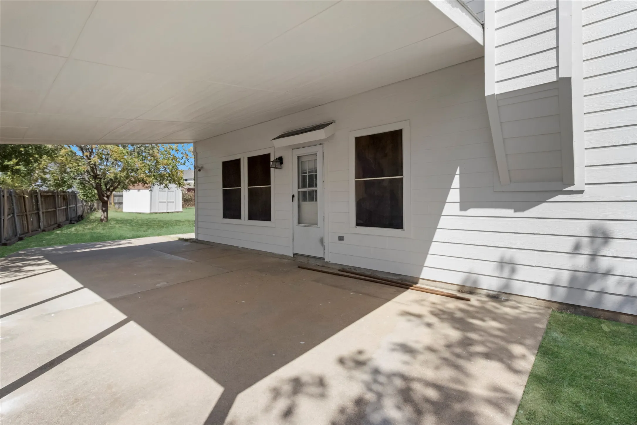 Covered concrete patio with a white paneled exterior, leading to a fenced yard with a detached shed