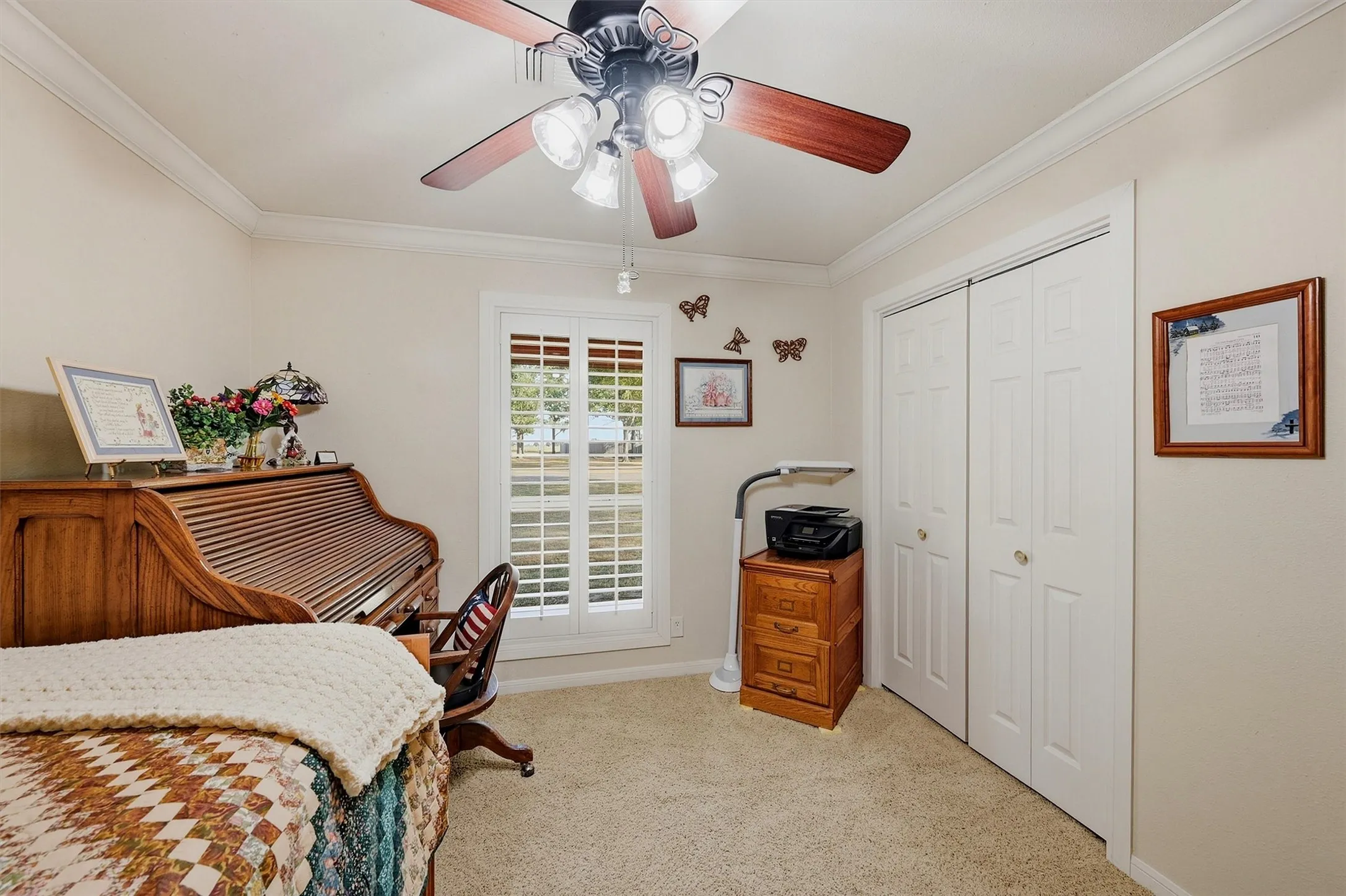 Window with plantation shutters, ceiling fan with light fixture, white bi-fold closet doors, crown molding, and carpeted flooring