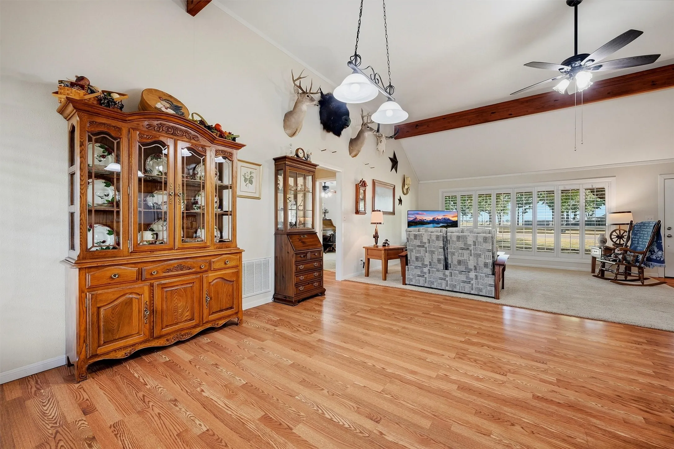 Vaulted ceiling with exposed wood beams, wood-finish flooring, ceiling fan, and large windows with plantation shutters