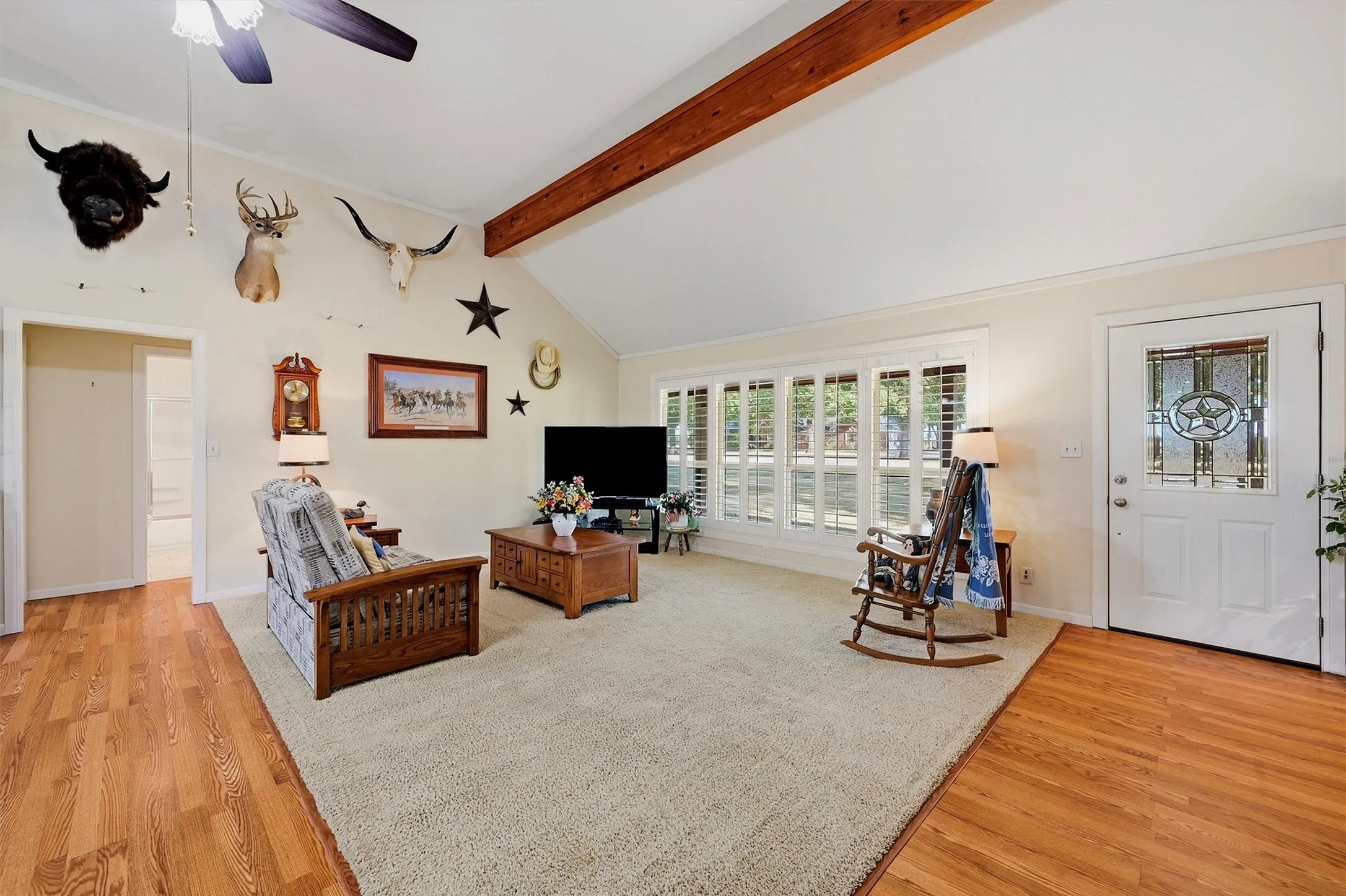 Vaulted ceiling with exposed wood beam, wood-finish flooring, entry door with decorative glass insert, and window shutters