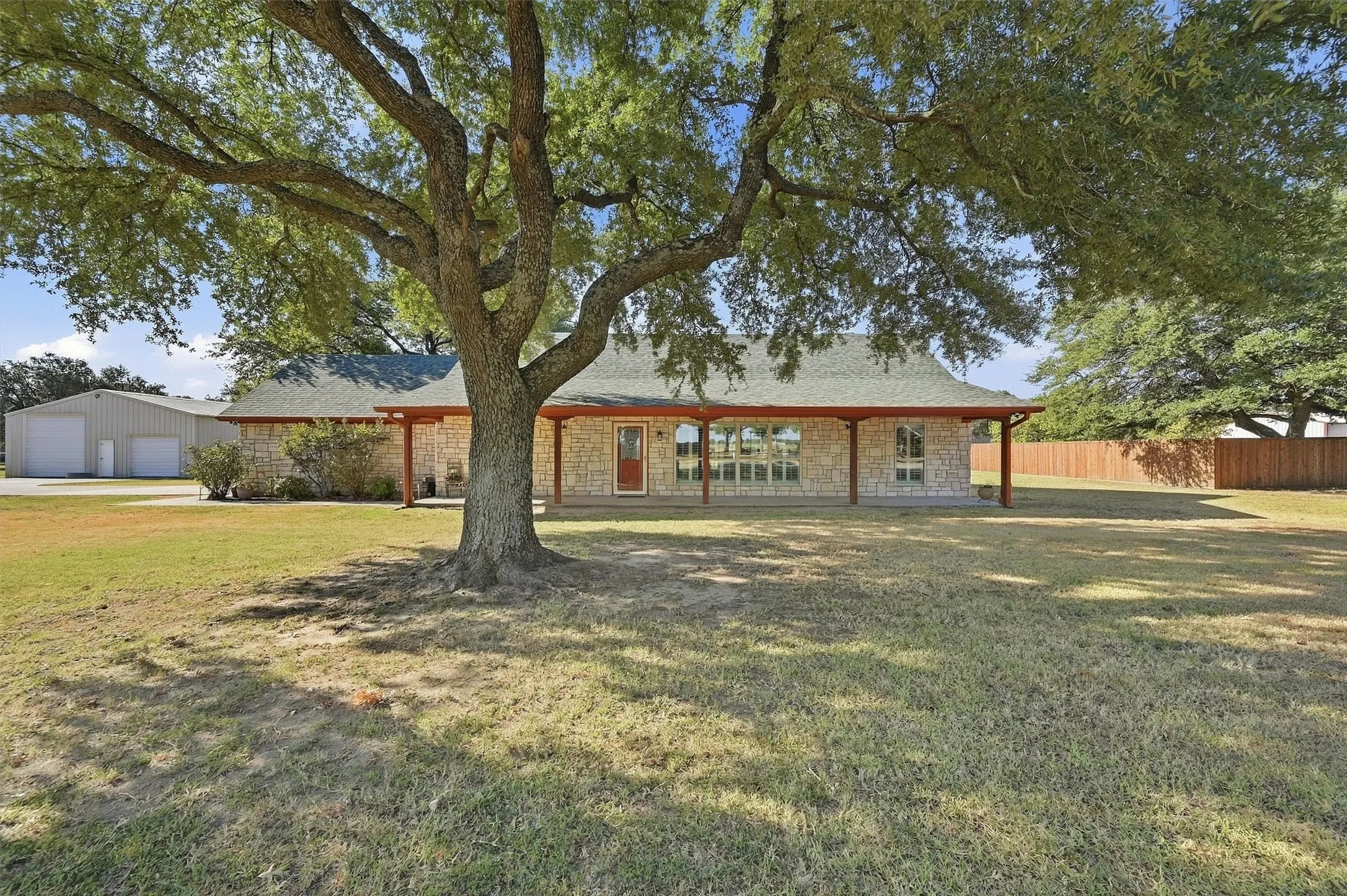 Stone facade, covered front porch, expansive lawn, mature shade tree, detached metal building with multiple garage doors