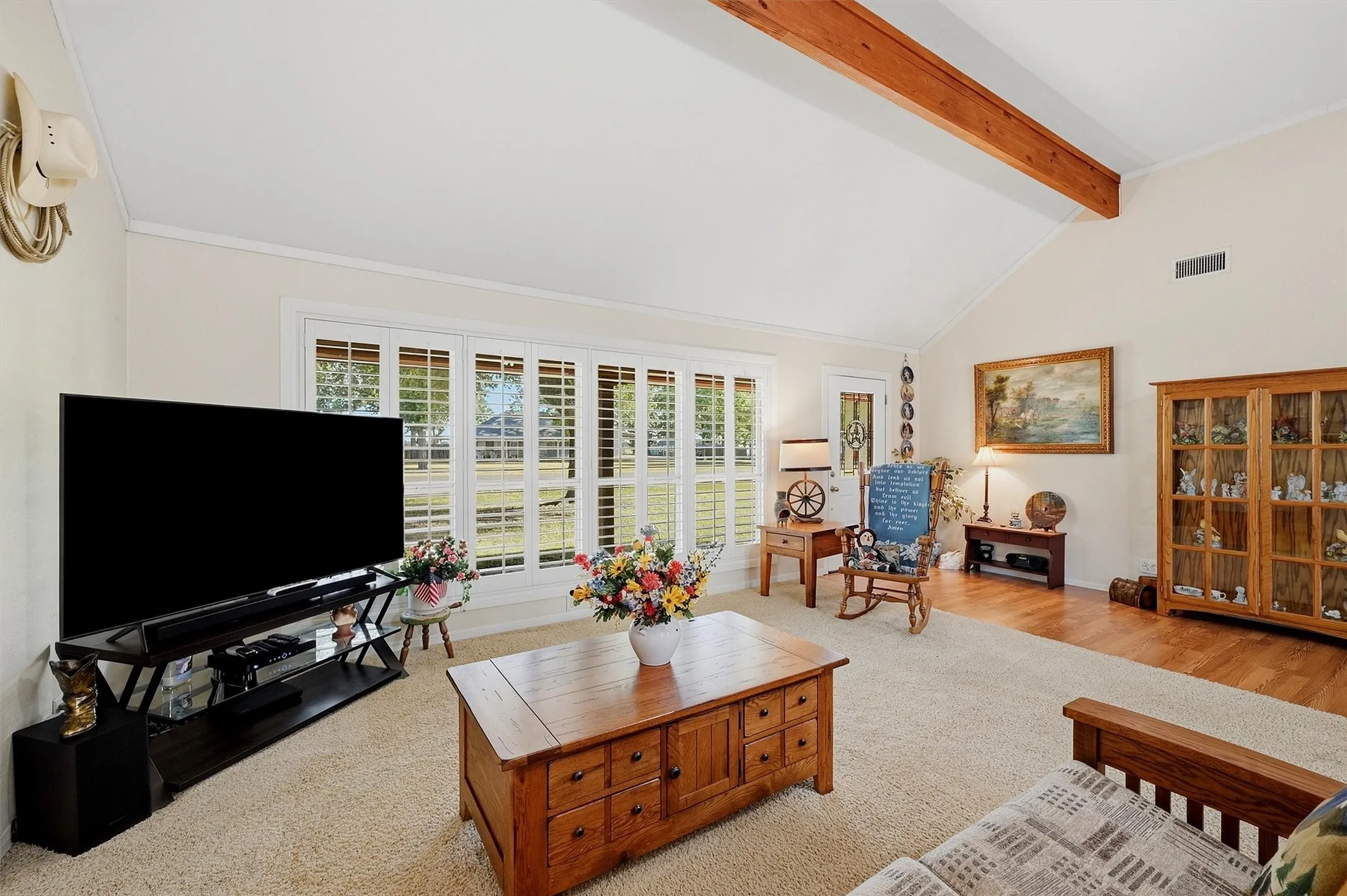 Vaulted ceiling with exposed wood beam, bay window with plantation shutters, light-toned wall paint, and mixed flooring of carpet and wood-finish