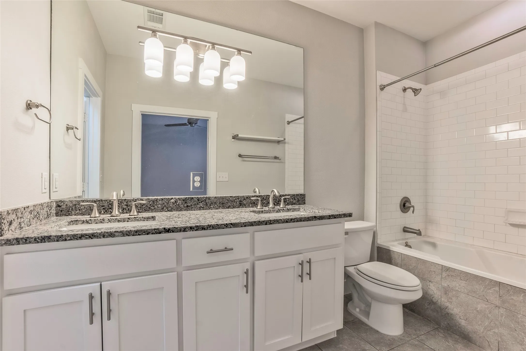 Dual vanity with stone countertop and undermount sinks, bright overhead lighting, white subway tile shower surround, and gray tiled flooring