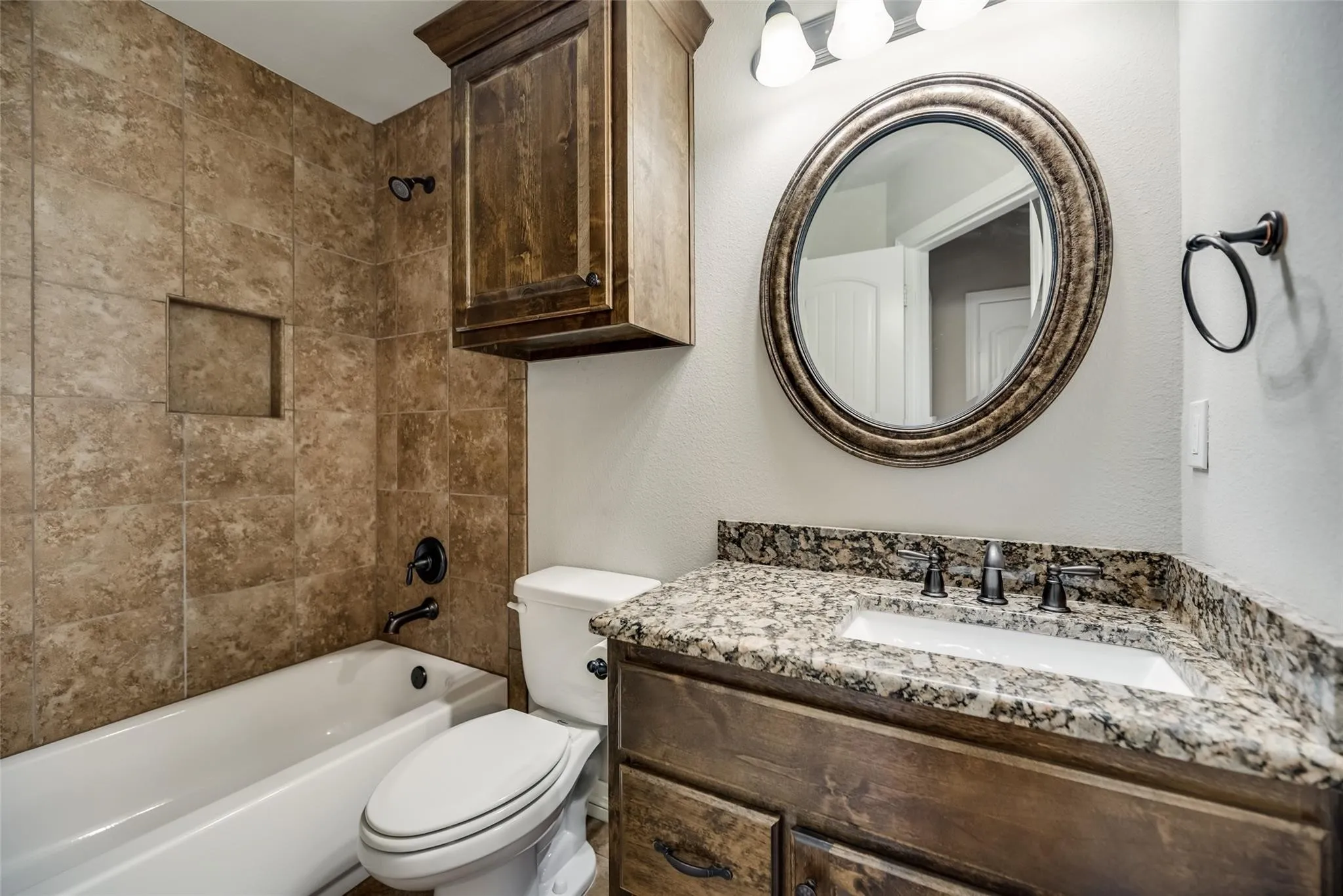 Granite-style countertop with an undermount sink, wood-finish vanity, tiled shower surround with recessed niche, and an oval framed mirror