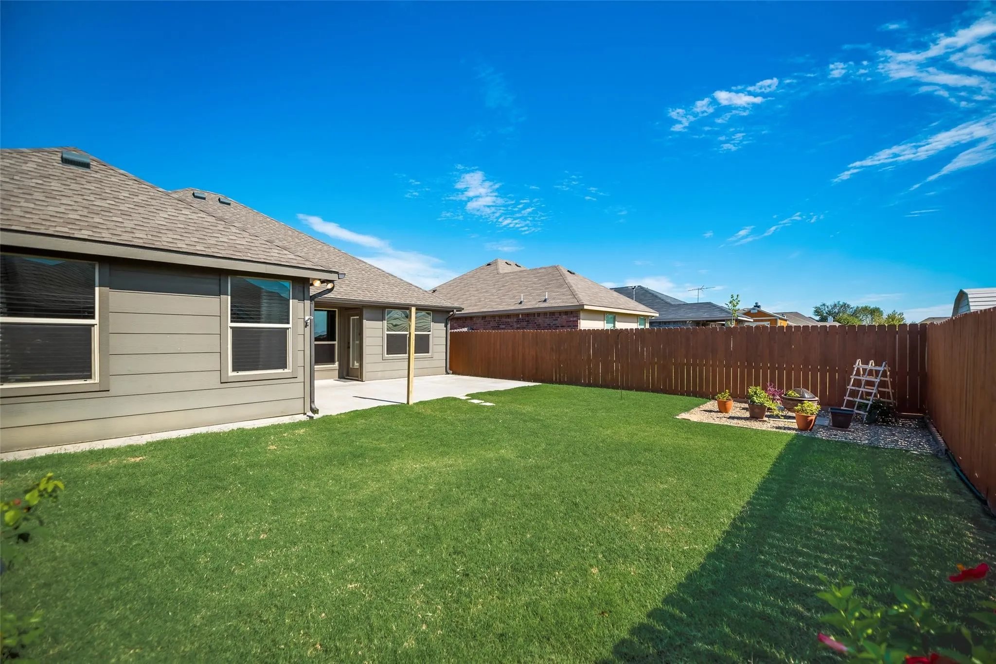 Expansive grassy yard with a wooden privacy fence, concrete patio, and a shingled roof