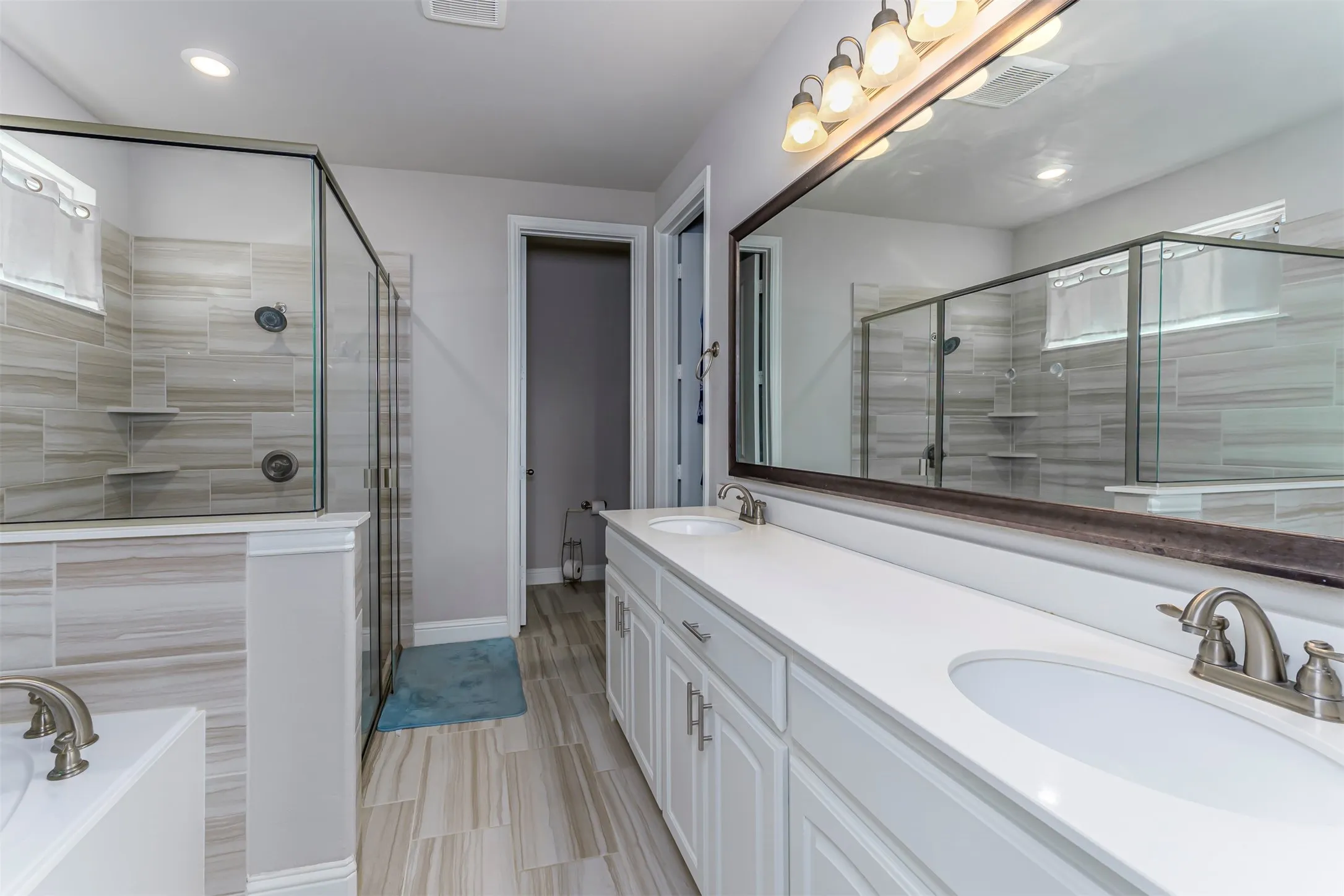 Dual vanity with white cabinetry and integrated sinks, a large framed mirror, a walk-in shower with clear glass enclosure and tiled walls, and wood-finish flooring