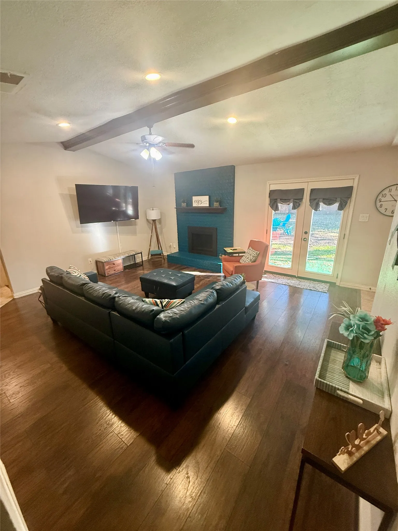 Recessed lighting, wood-finish flooring, exposed ceiling beams, fireplace with a teal-painted surround, and double French doors to the exterior