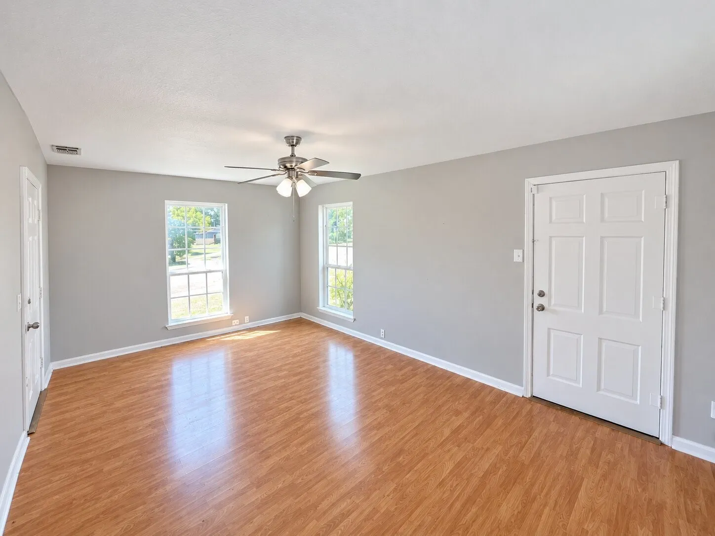 Wood-finish flooring, two large windows with grid patterns, ceiling fan with integrated lighting, and a paneled entry door