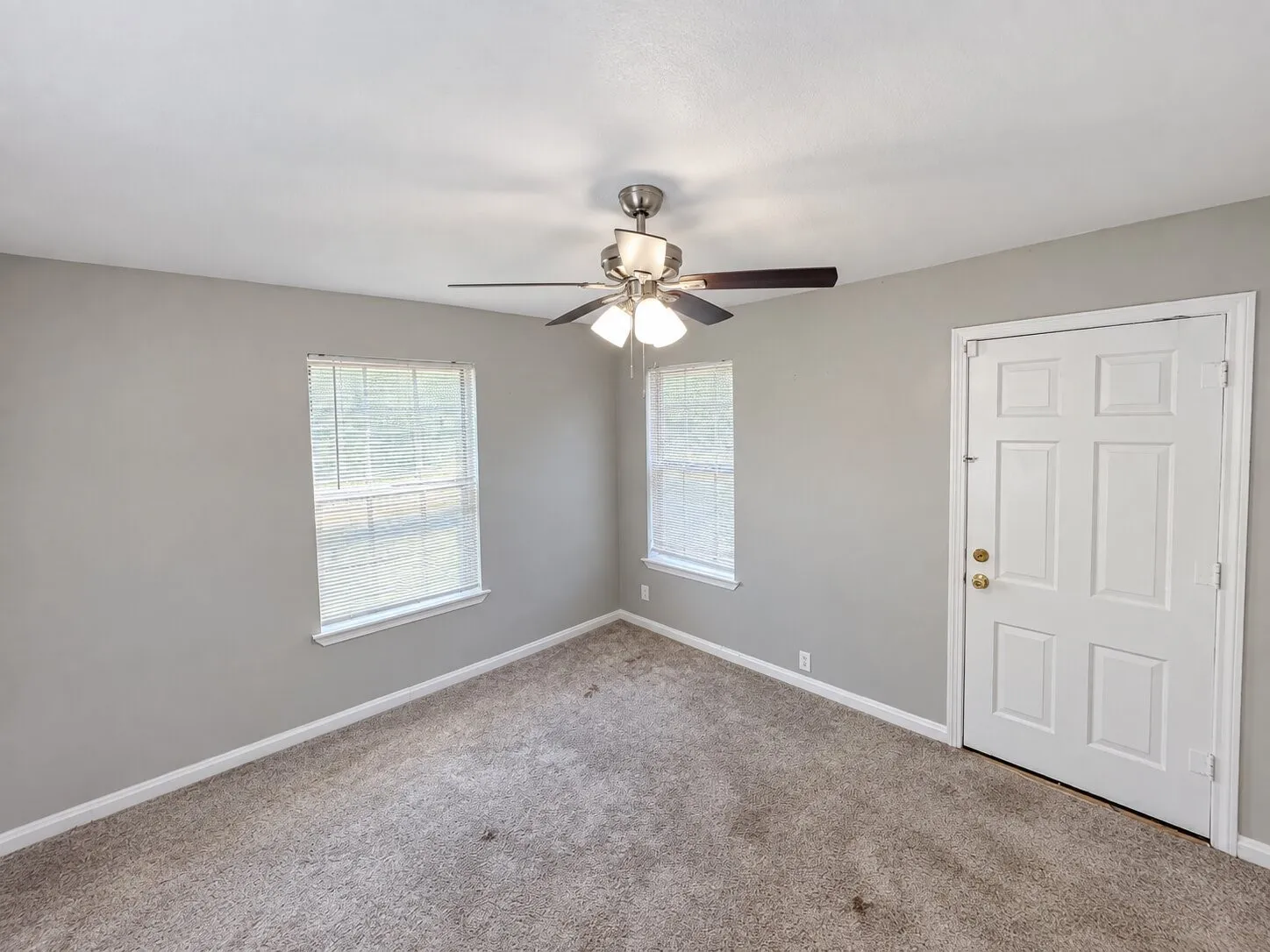 Neutral gray wall paint, tan carpet, two windows with blinds, a white paneled door with brass hardware, and a ceiling fan with light fixture