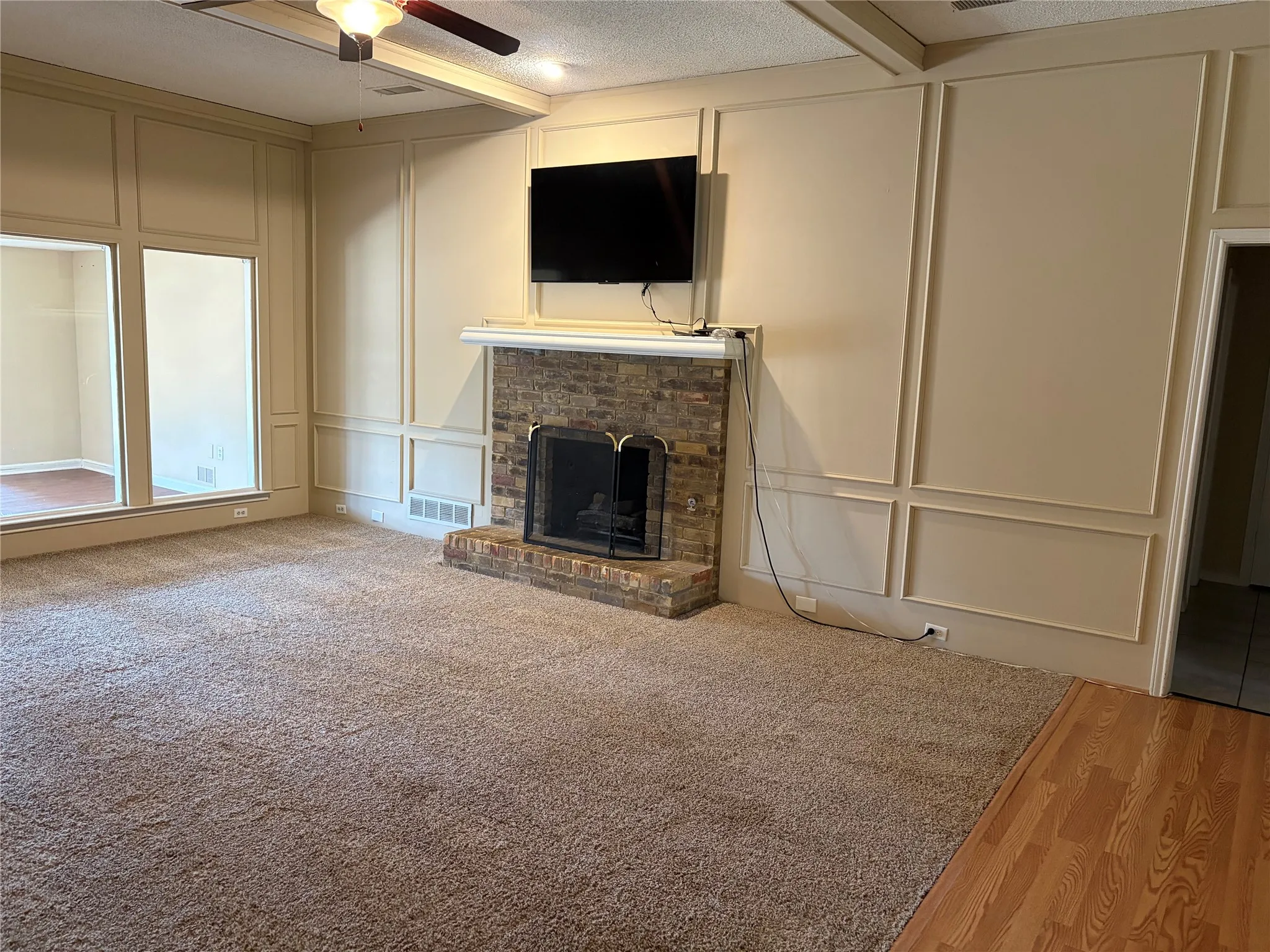 Stone-faced fireplace with a white mantel, large picture windows, decorative wall paneling, ceiling fan with light fixture, and wood-finish flooring