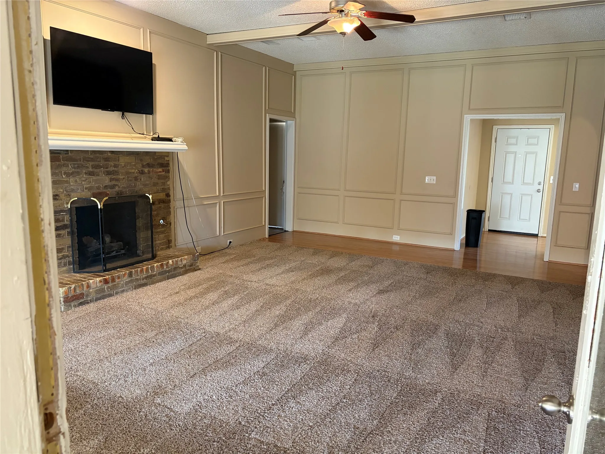 Living area featuring a brick fireplace with a white mantel, decorative wall paneling, a ceiling fan with integrated lighting, and a mix of carpeted and wood-finish flooring