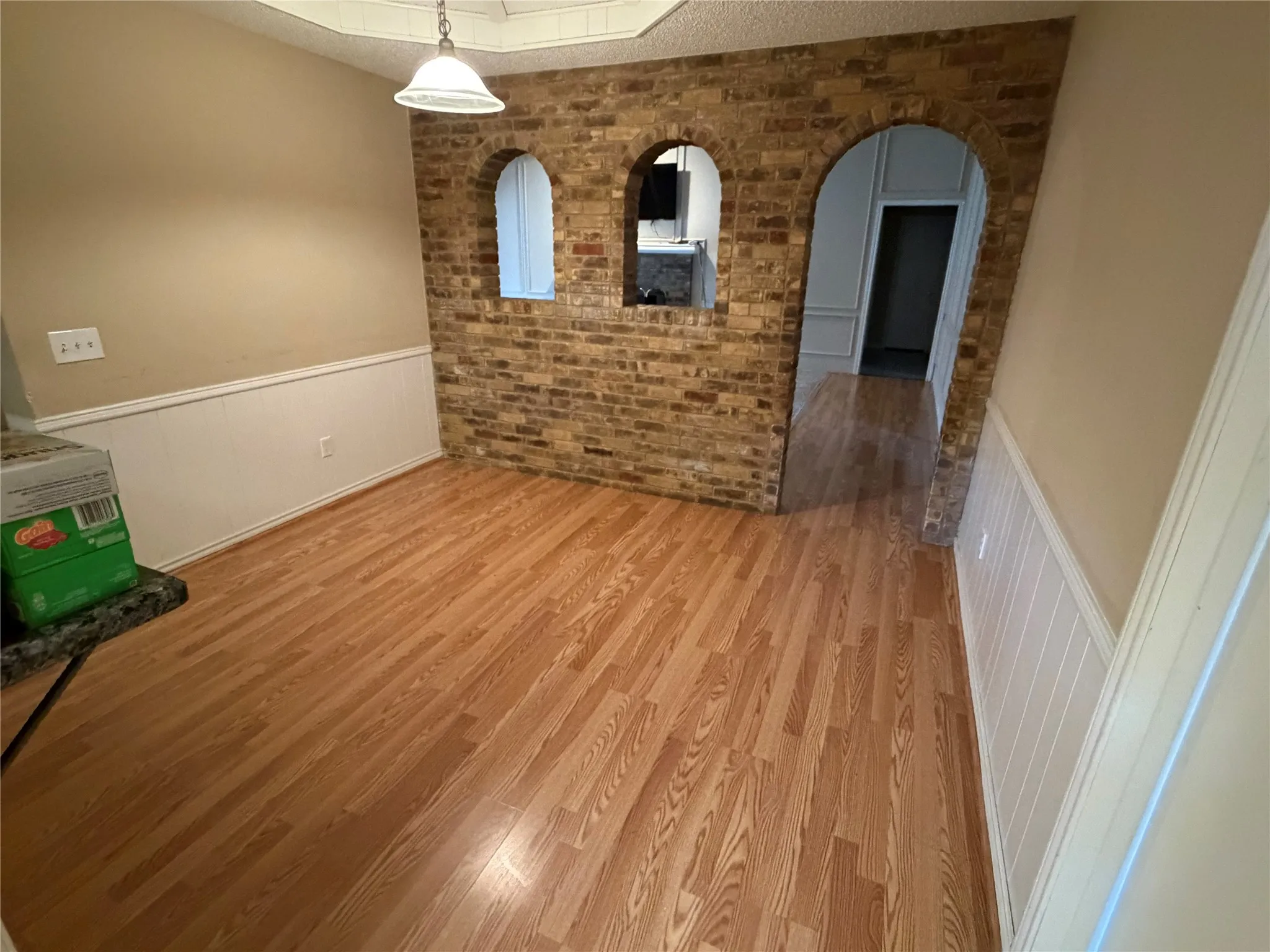 Hardwood-finish flooring, brick accent wall with arched openings, white paneled wainscoting, and a built-in ceiling light fixture
