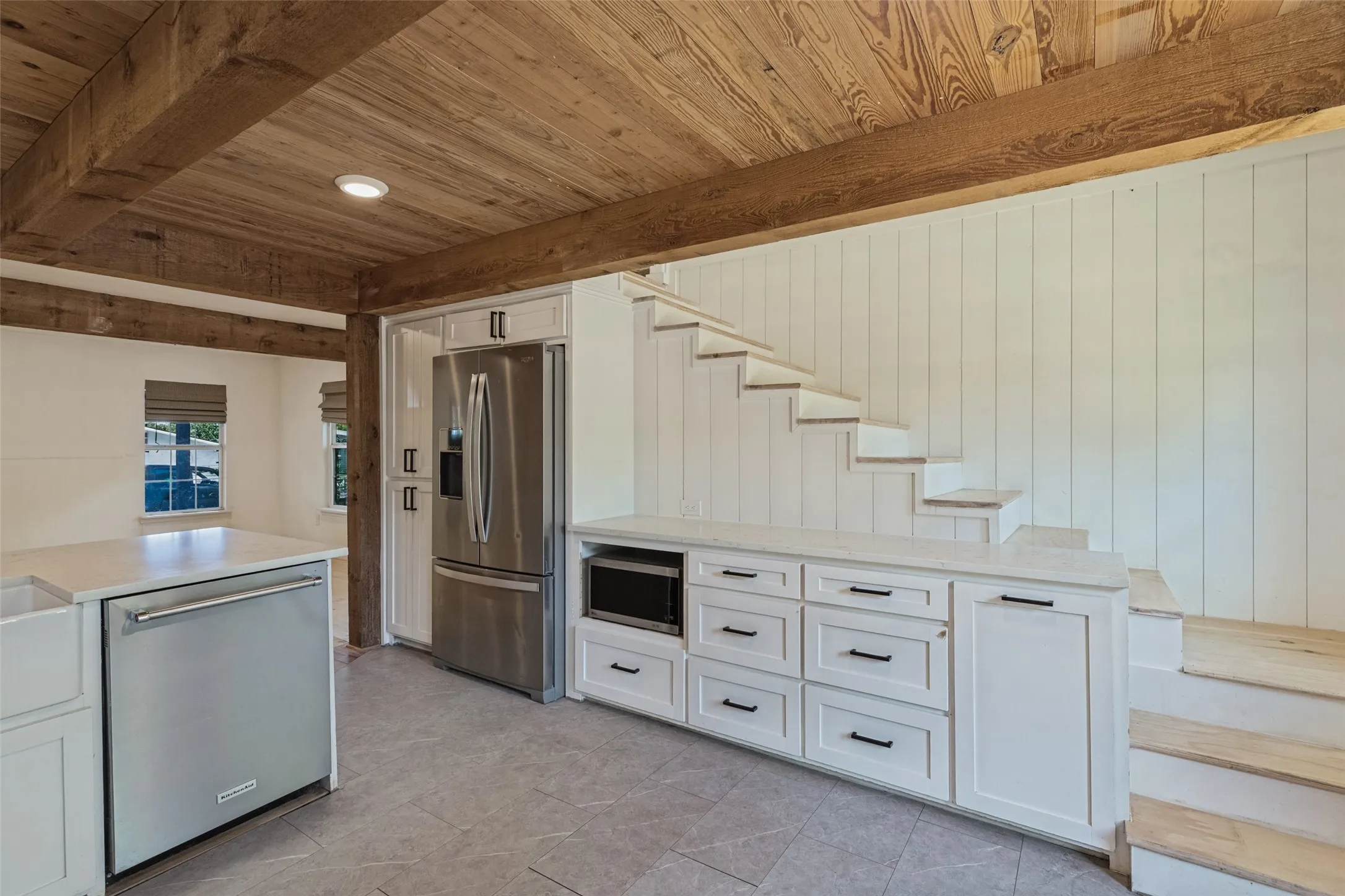 Wood-paneled ceiling with exposed beams, white cabinetry, integrated stainless steel appliances, light-toned flooring, and open-tread staircase