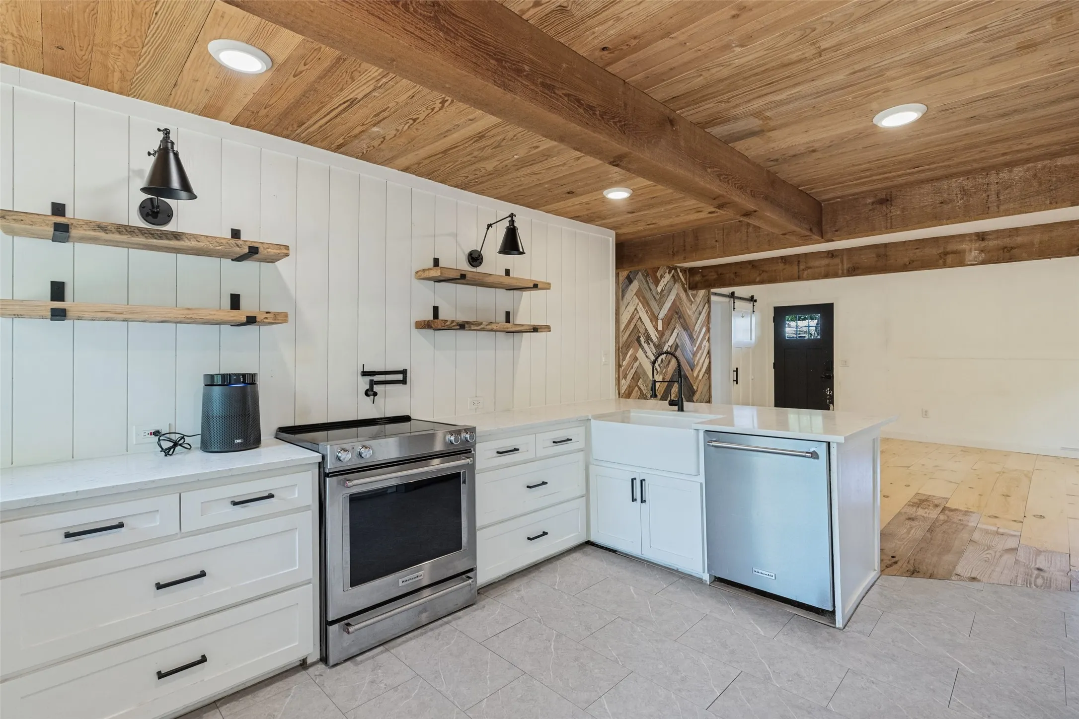 Exposed wood beam ceiling, white shaker cabinetry with dark hardware, stainless steel appliances, farmhouse sink with gooseneck faucet, and light tile flooring