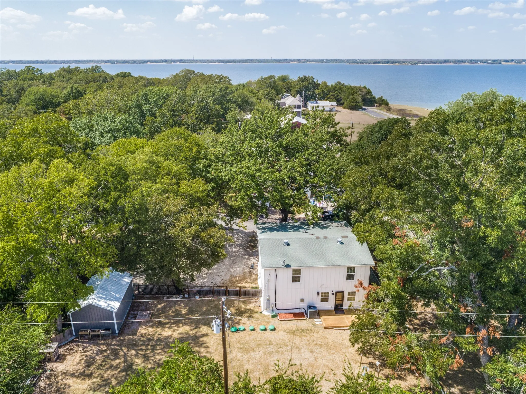 Waterfront property featuring extensive mature tree coverage, a two-story residence with a light-colored exterior and green roof, a detached shed with a light-colored roof, and a wooden deck