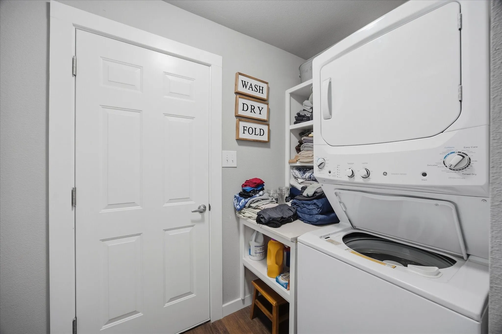 Laundry area featuring wood-finish flooring, white paneled door, built-in shelving, and a stacked laundry unit